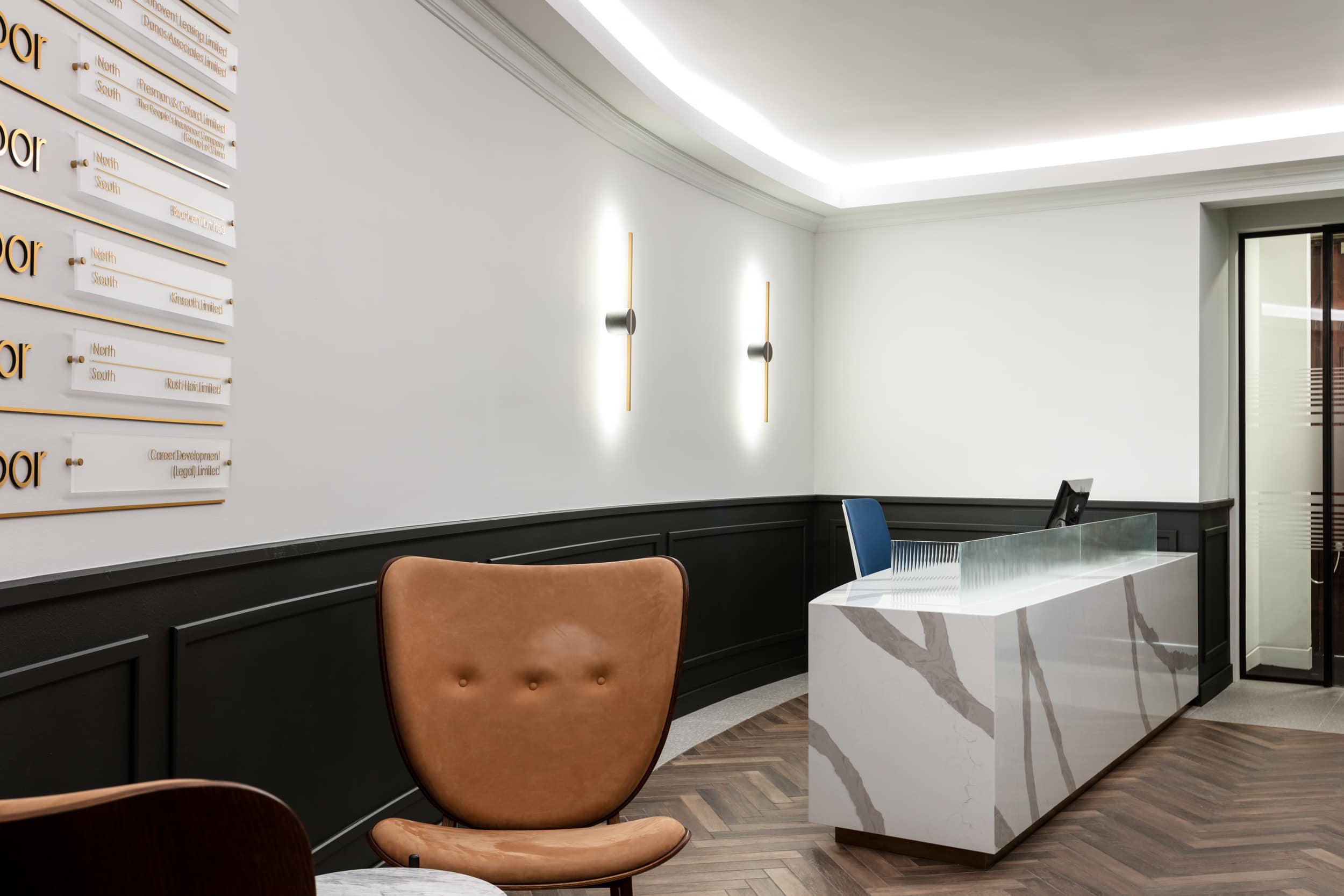 Reception area featuring a modern desk with a glass top, a brown upholstered chair, and wall-mounted lights, set against a dark paneled wall.
