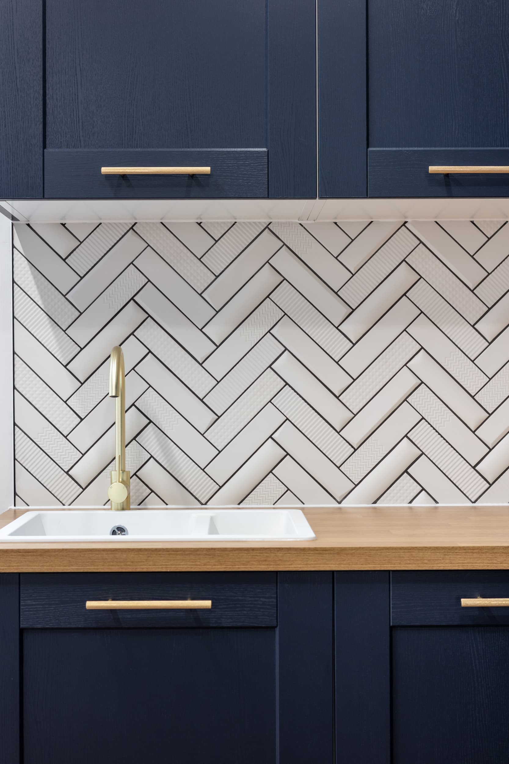 Modern kitchen featuring navy blue cabinets, a white herringbone tile backsplash, a gold faucet, and a white sink on a wooden countertop.