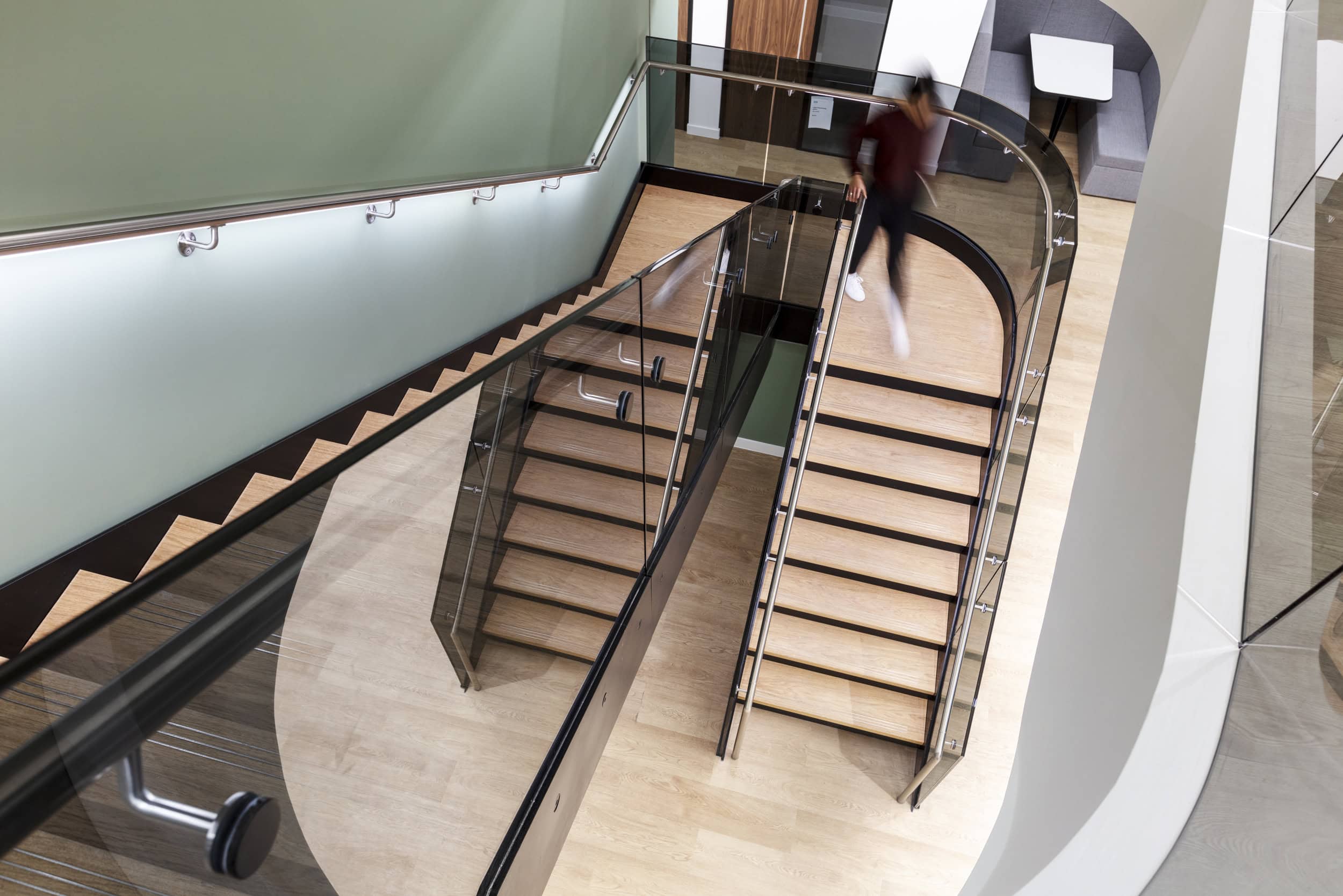 A modern staircase with wooden steps and glass railings, viewed from above, with a person walking down.