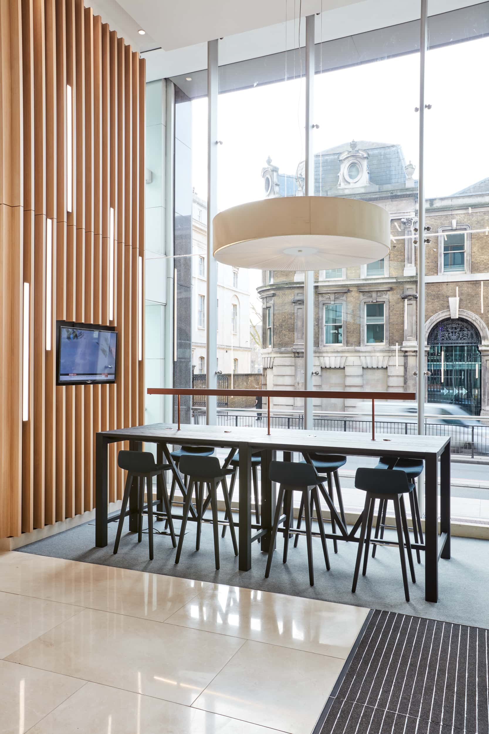 Modern interior featuring a wooden wall, a large circular light fixture, a high table with black stools, and a view of a historic building through large glass w