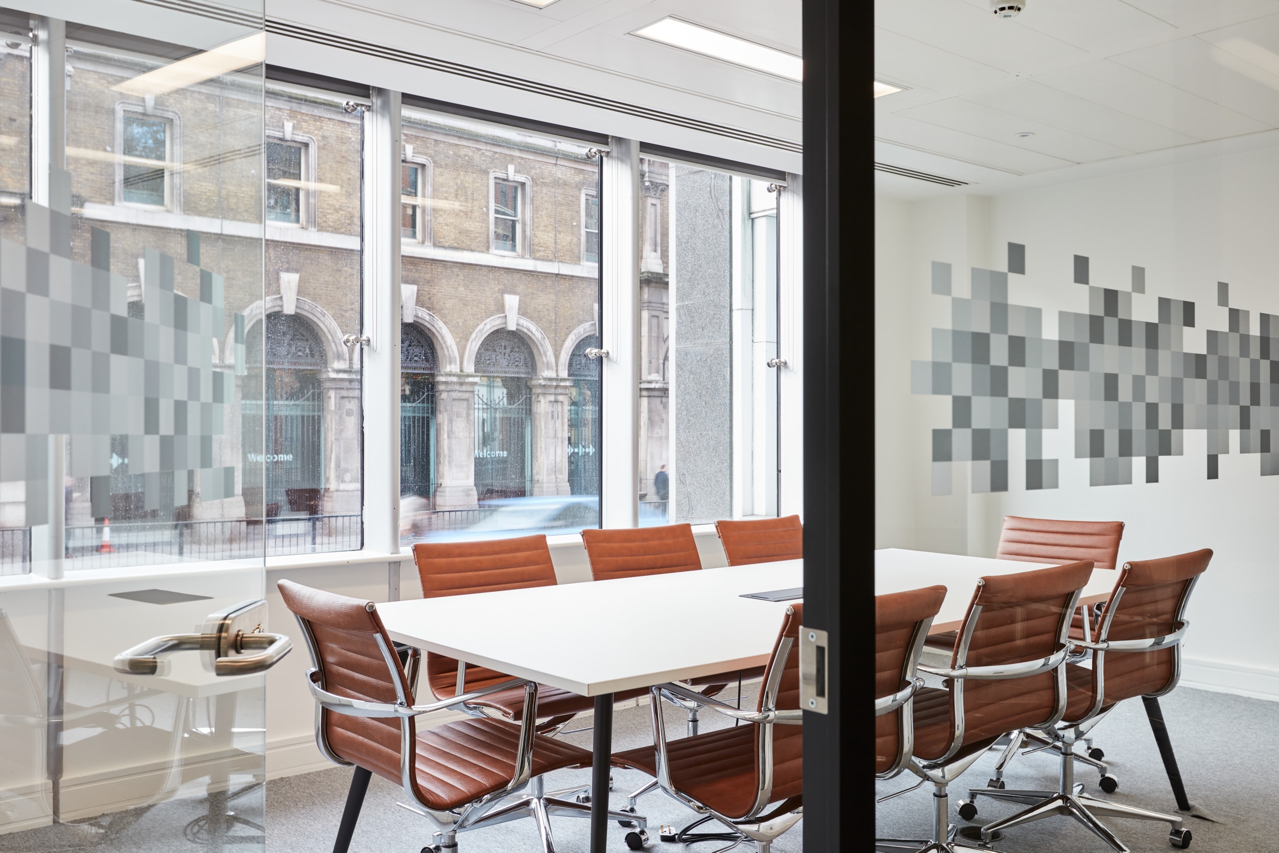 Conference room featuring a white table and brown leather chairs, with large windows displaying a view of a brick building and a "Welcome" sign.