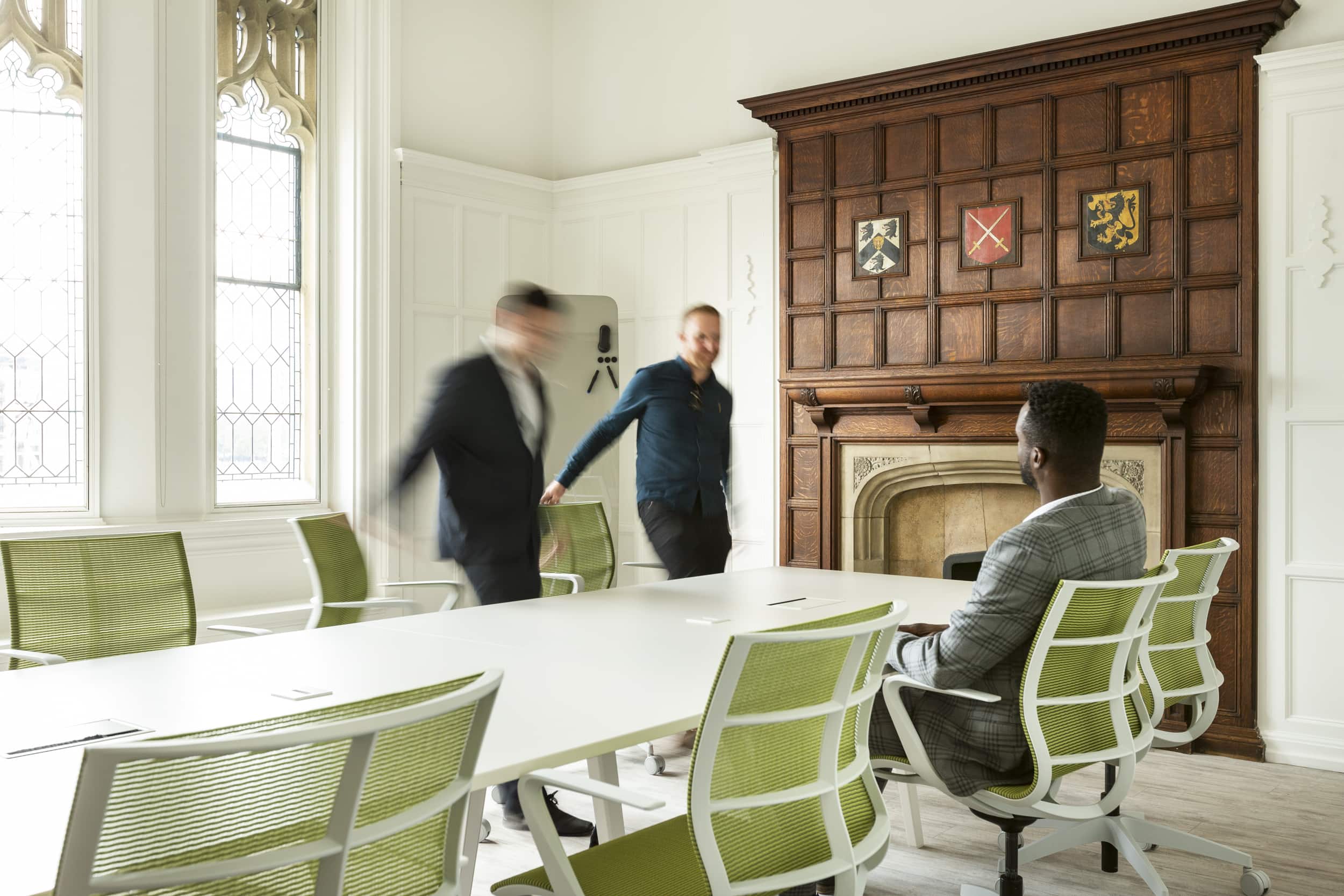 A meeting room with a long white table and green chairs, featuring a man in a suit sitting at the table while two blurred figures walk past a wooden fireplace a