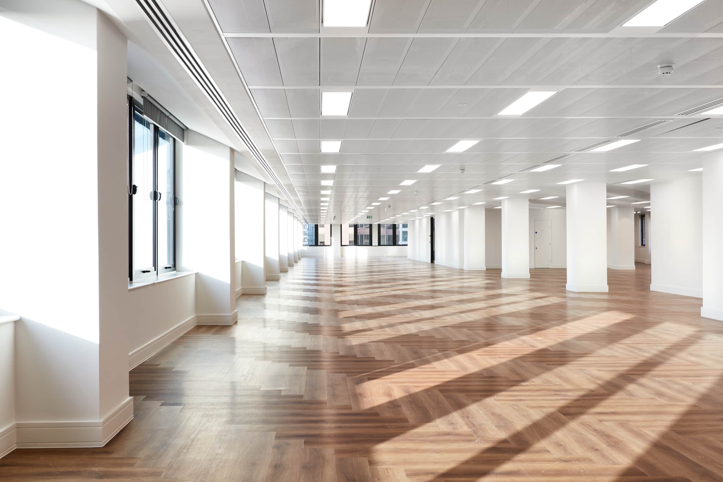 Spacious interior of an office with wooden flooring, large windows allowing natural light, and a modern ceiling design.