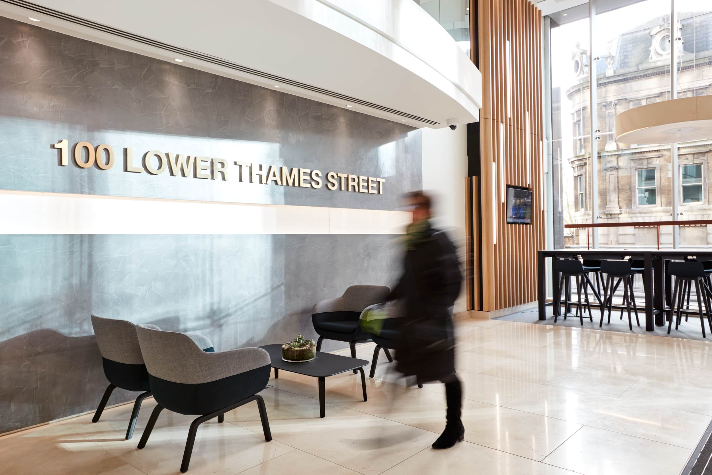 Reception area of 100 Lower Thames Street featuring modern seating, a coffee table, and a large wall sign, with a blurred figure walking through.