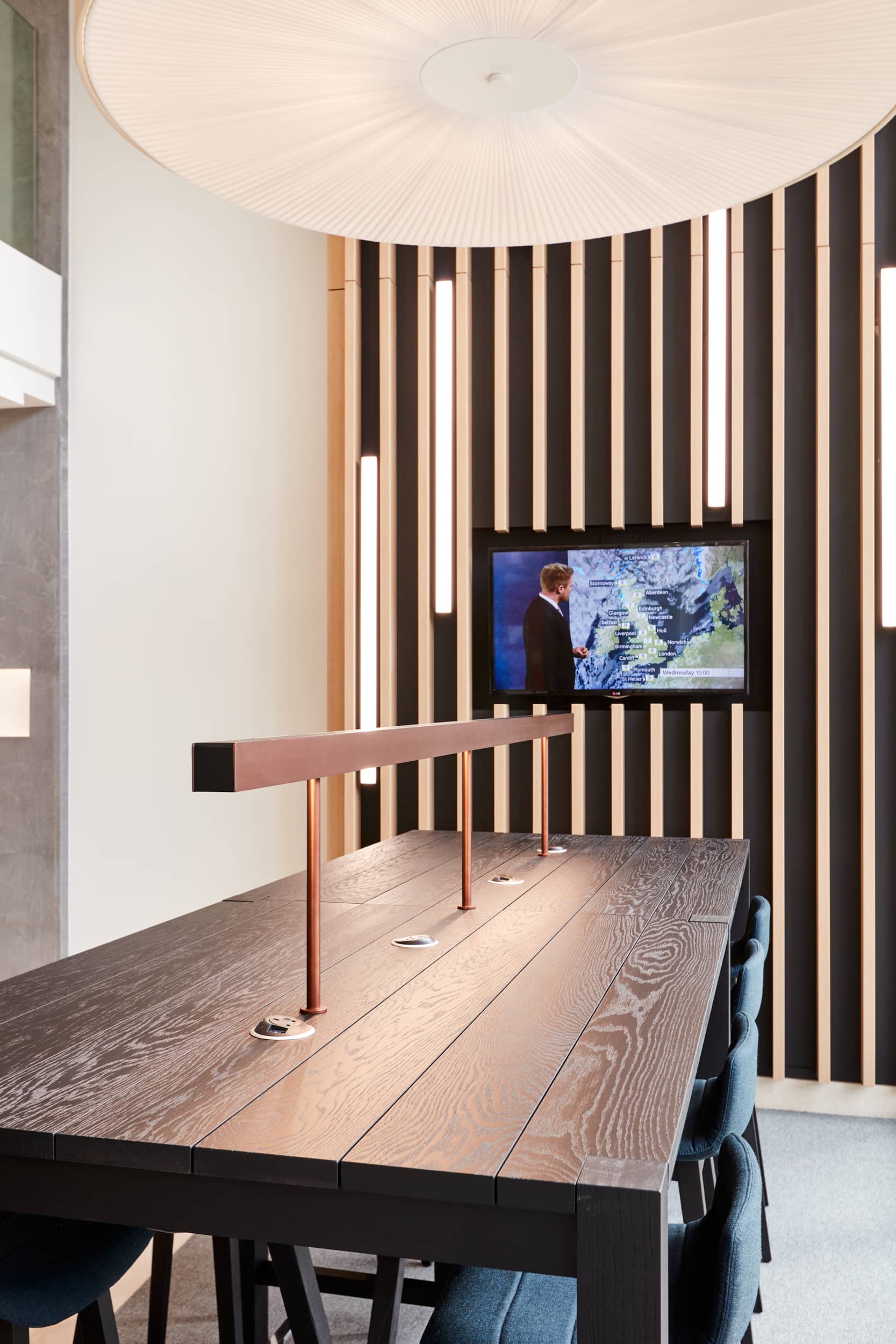 Modern meeting room featuring a dark wooden table with built-in power outlets, blue chairs, and a wall-mounted screen displaying a weather report, surrounded by