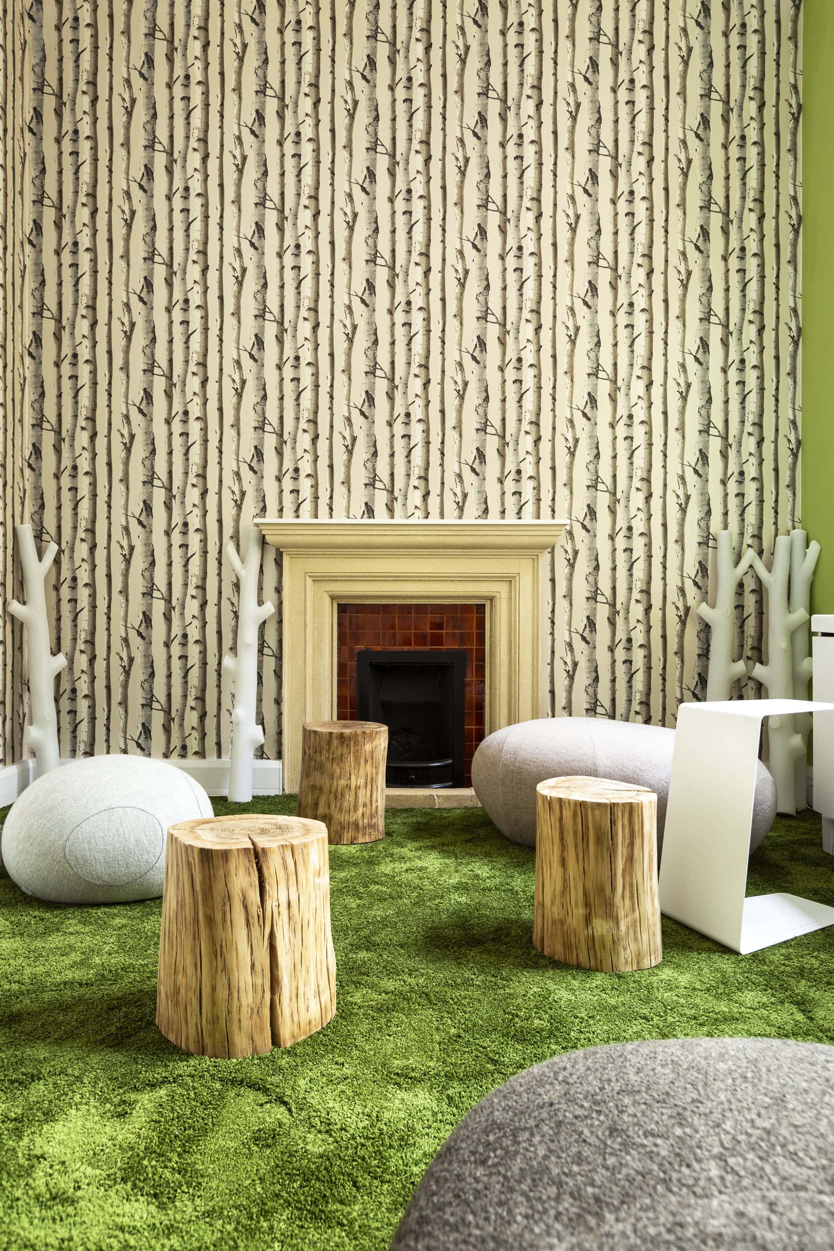 Interior of Allianz Sion Hall featuring a green carpet, wooden stools, soft seating, a decorative fireplace, and birch tree-patterned wallpaper.