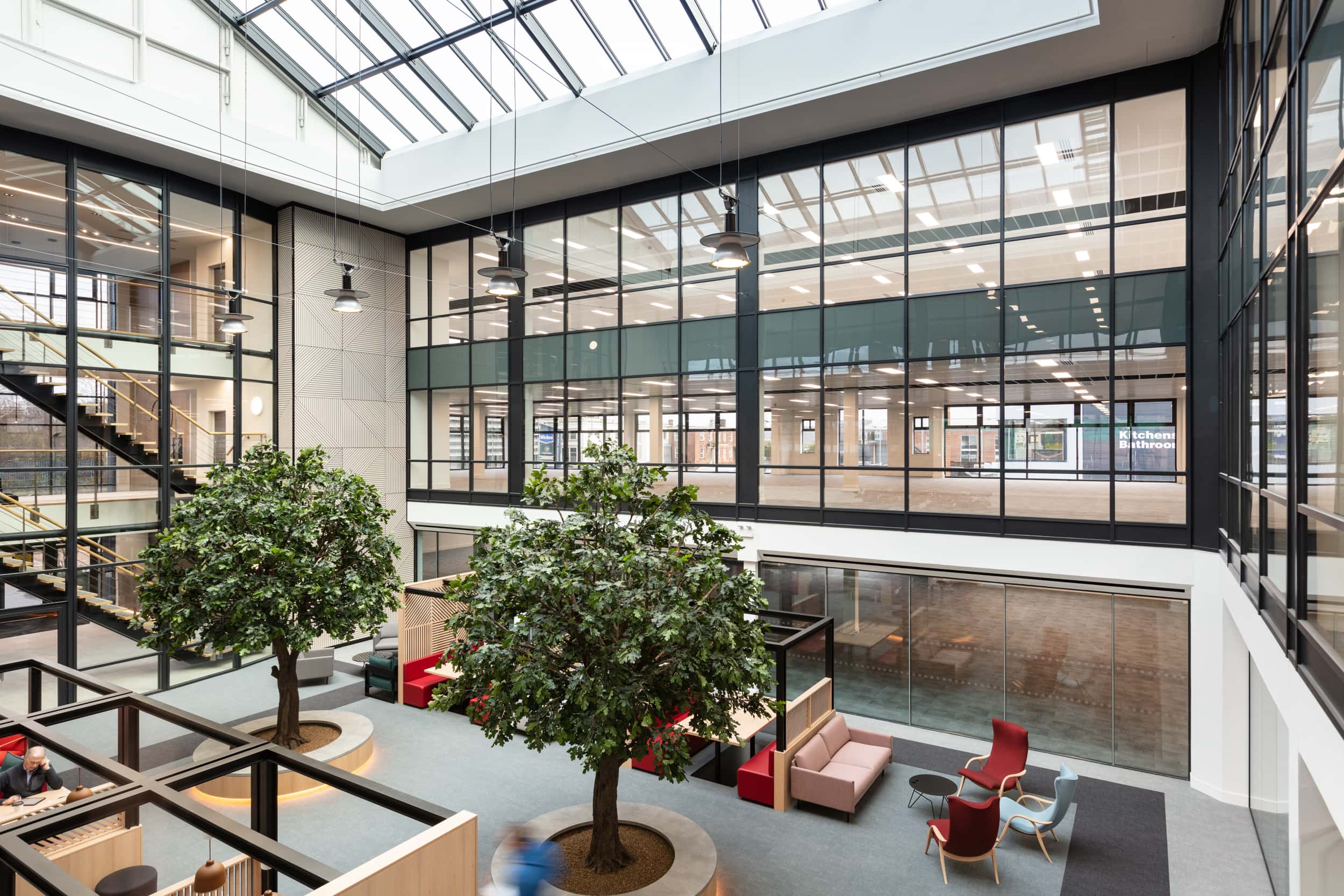 Interior view of a modern atrium featuring large glass windows, two indoor trees, and seating areas with red and blue chairs.