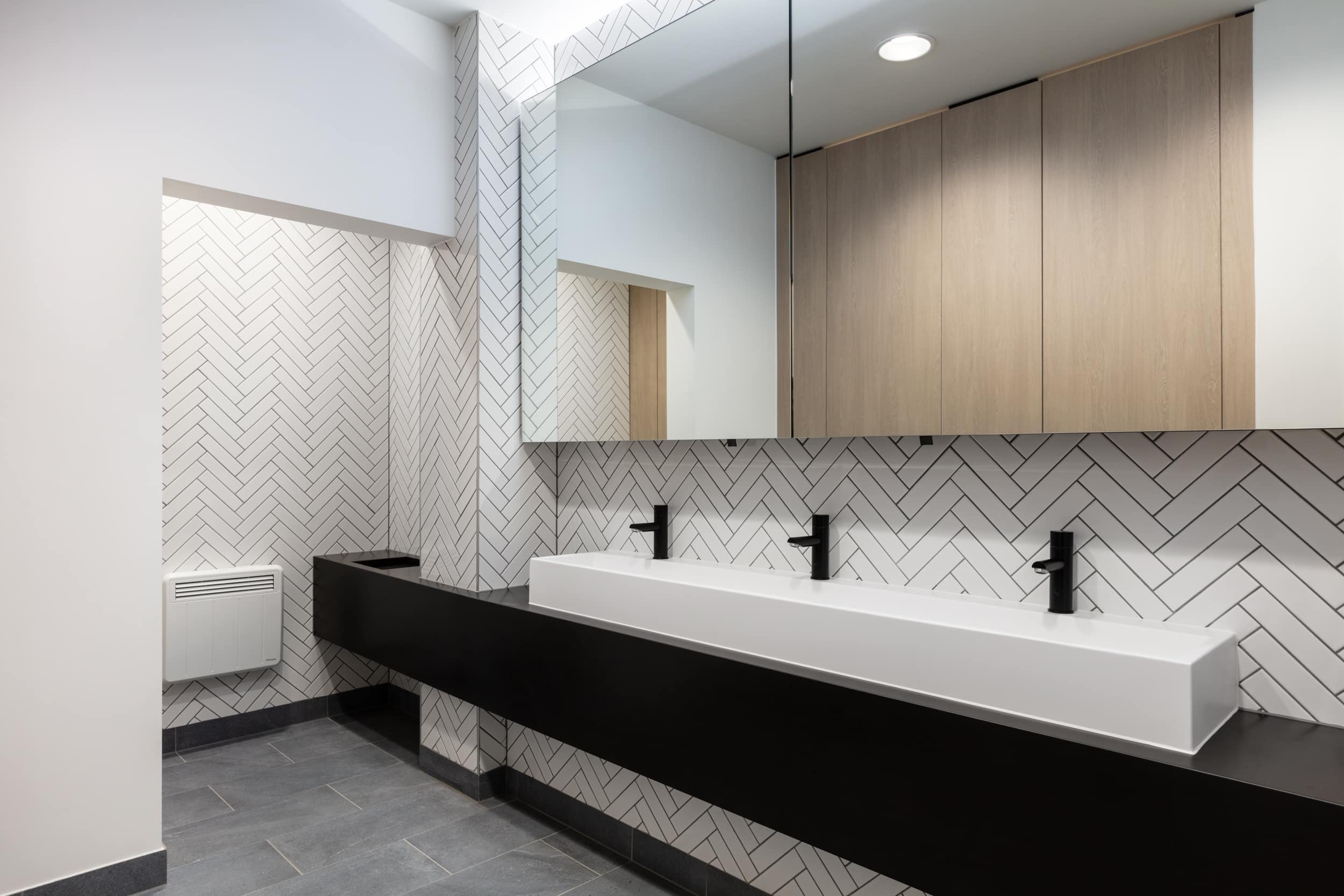Modern bathroom featuring a long white sink with black faucets, herringbone-patterned tile walls, and wooden cabinetry above a black countertop.