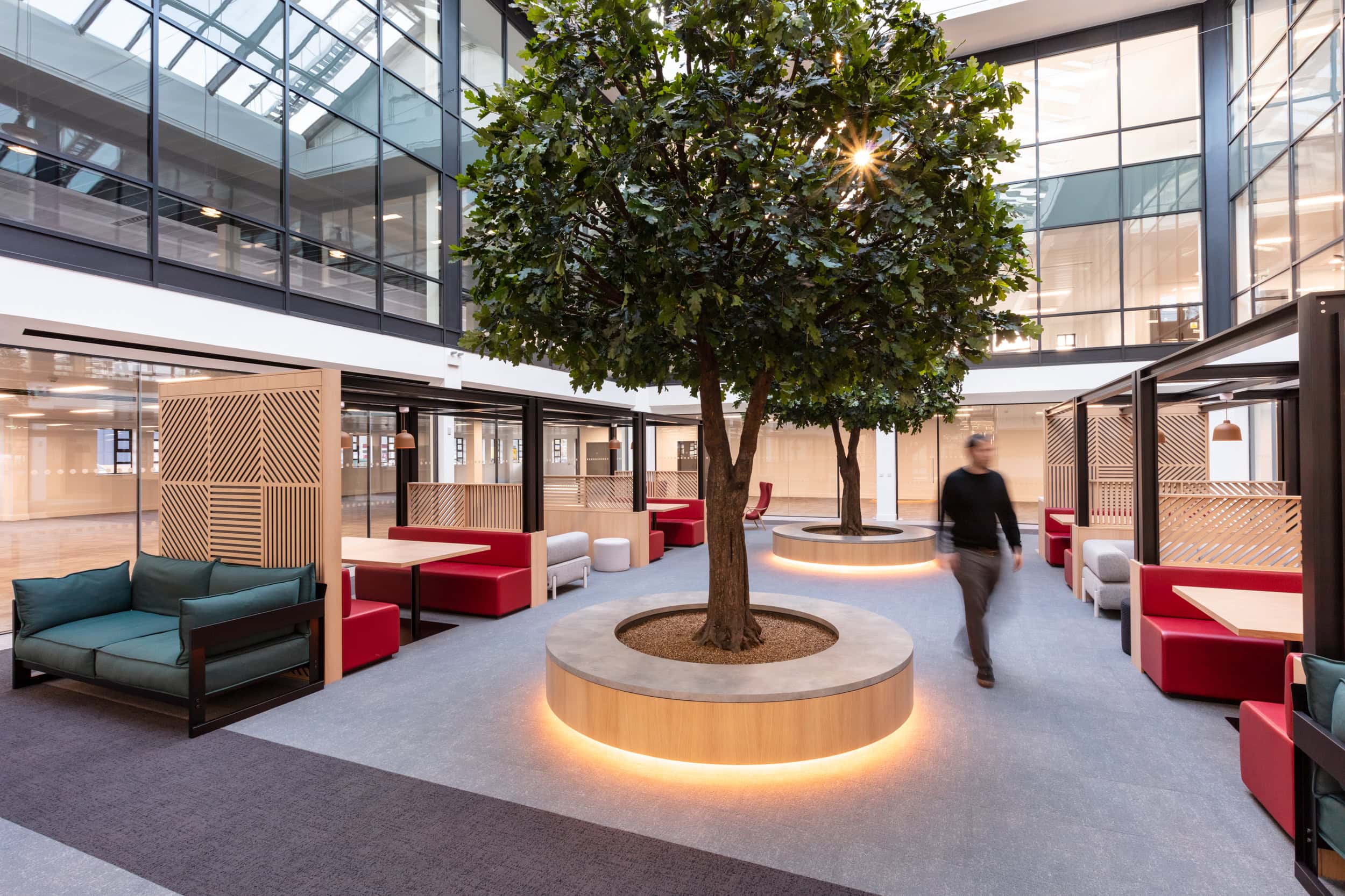 Modern indoor workspace featuring a large artificial tree, seating areas with red and green couches, and glass walls allowing natural light.