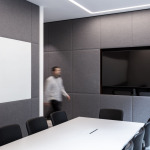 Modern conference room featuring a large whiteboard, a wall-mounted television, and a long table surrounded by black chairs, with a person walking in the backgr
