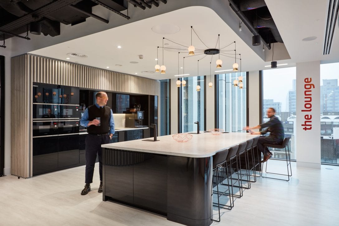 Two employees are shown inside an office tea point. Above the kitchen island bench there is a feature pendant light that is illuminated with warm lights. 