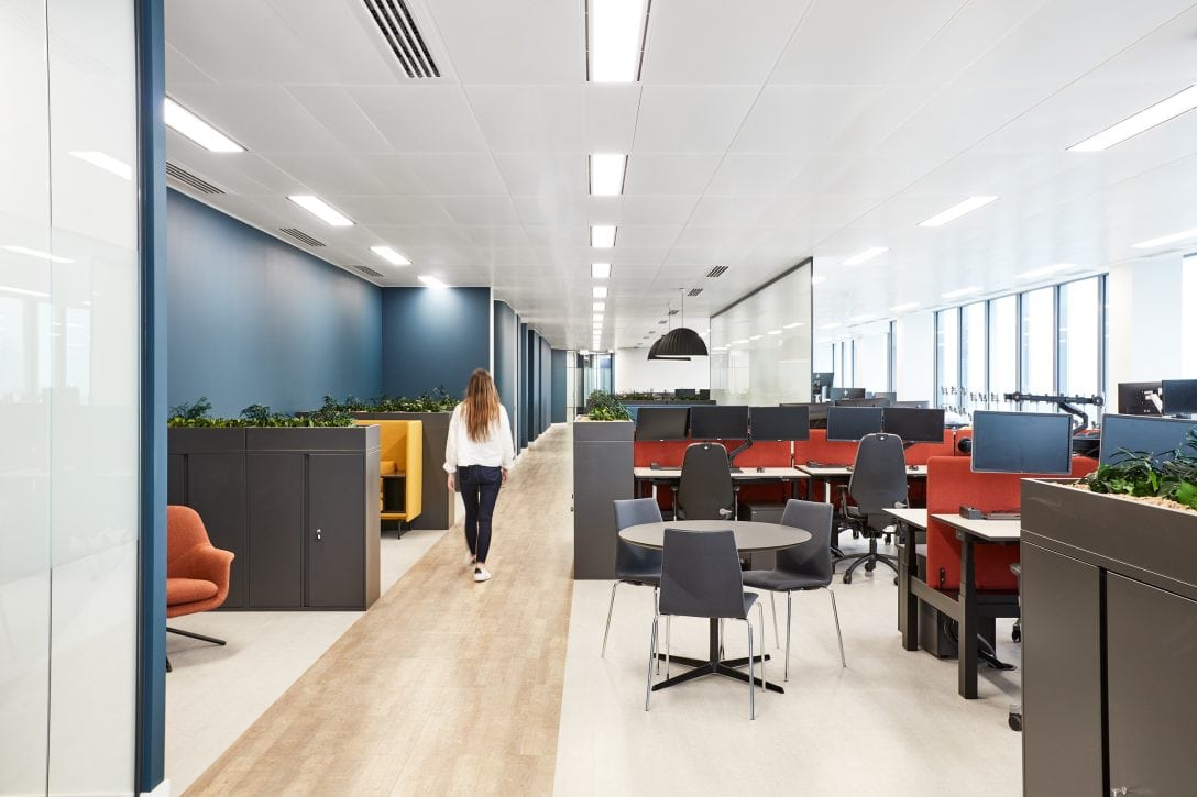 A girl is shown walking down a corridor inside an office, there are desks to the right and collaboration zones with different furniture configurations are on offer to the left.