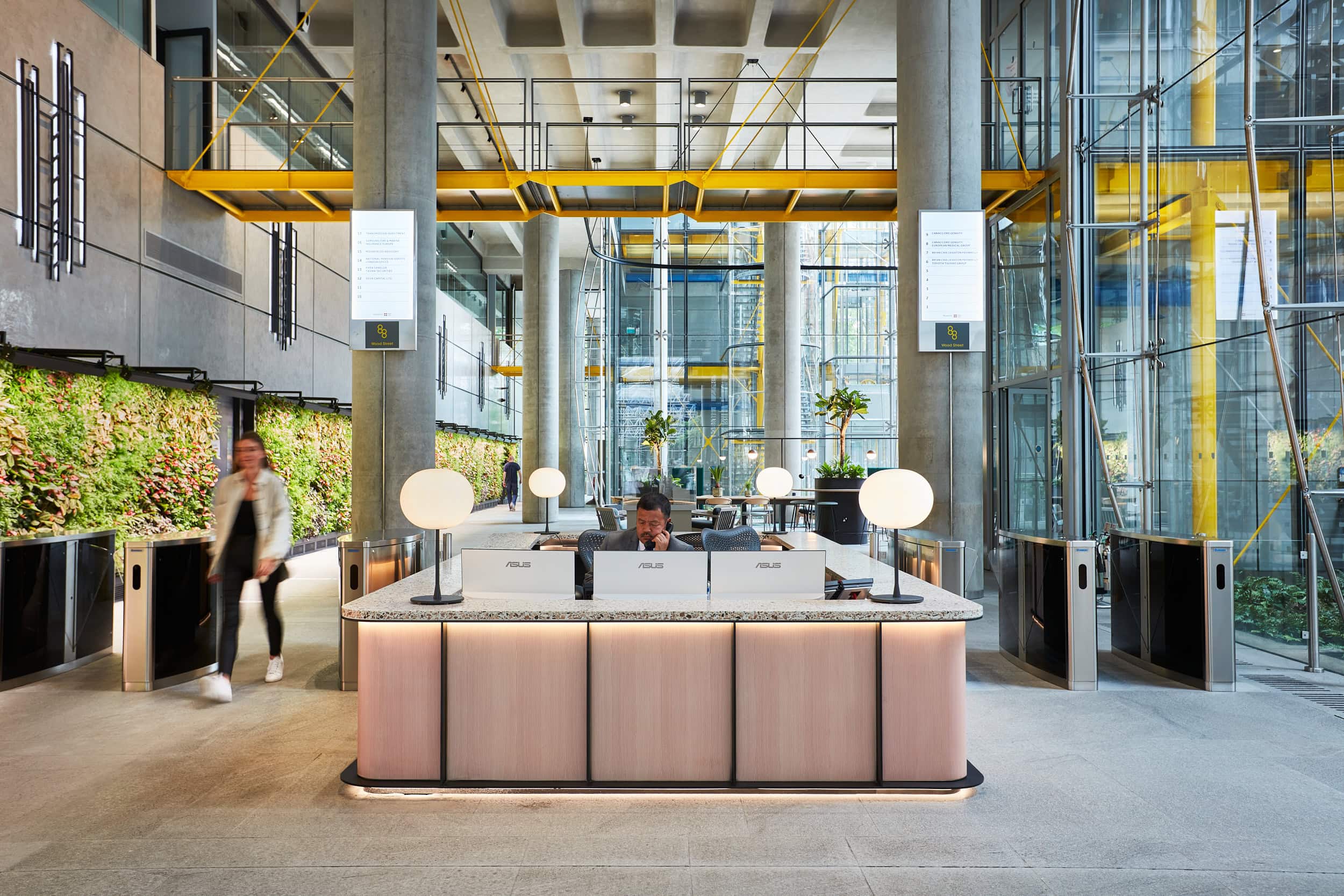 Modern office reception area featuring a sleek front desk with round lamps, surrounded by greenery and large glass windows, with a person walking past.