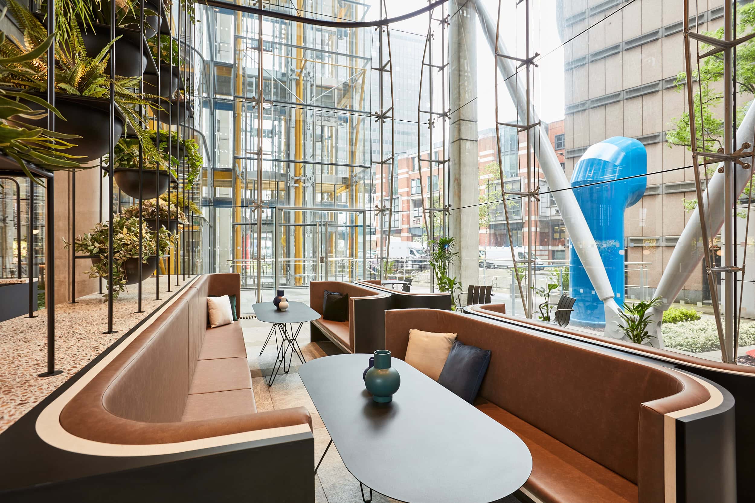 Modern reception waiting area featuring curved brown seating, black tables, and hanging plants, with large glass windows revealing an urban view.