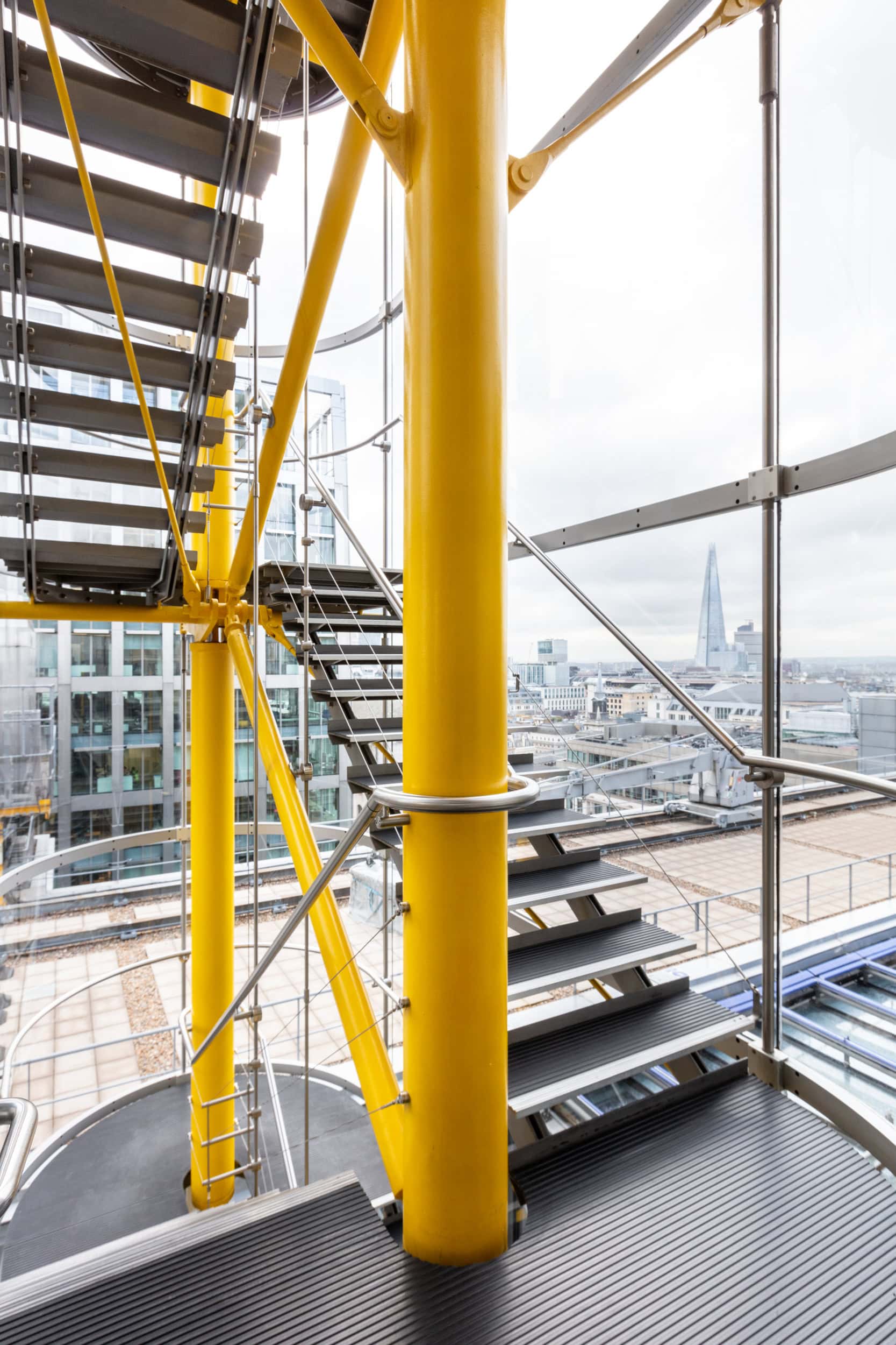 Interior view of a modern staircase with yellow support columns and metal railings, overlooking a cityscape through large glass windows.