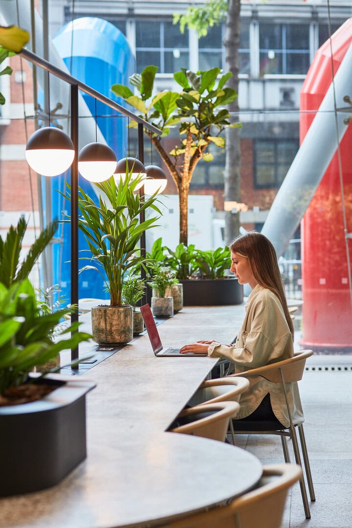 A woman working on a laptop at a modern workspace with plants and decorative lighting, featuring colorful architectural elements in the background.
