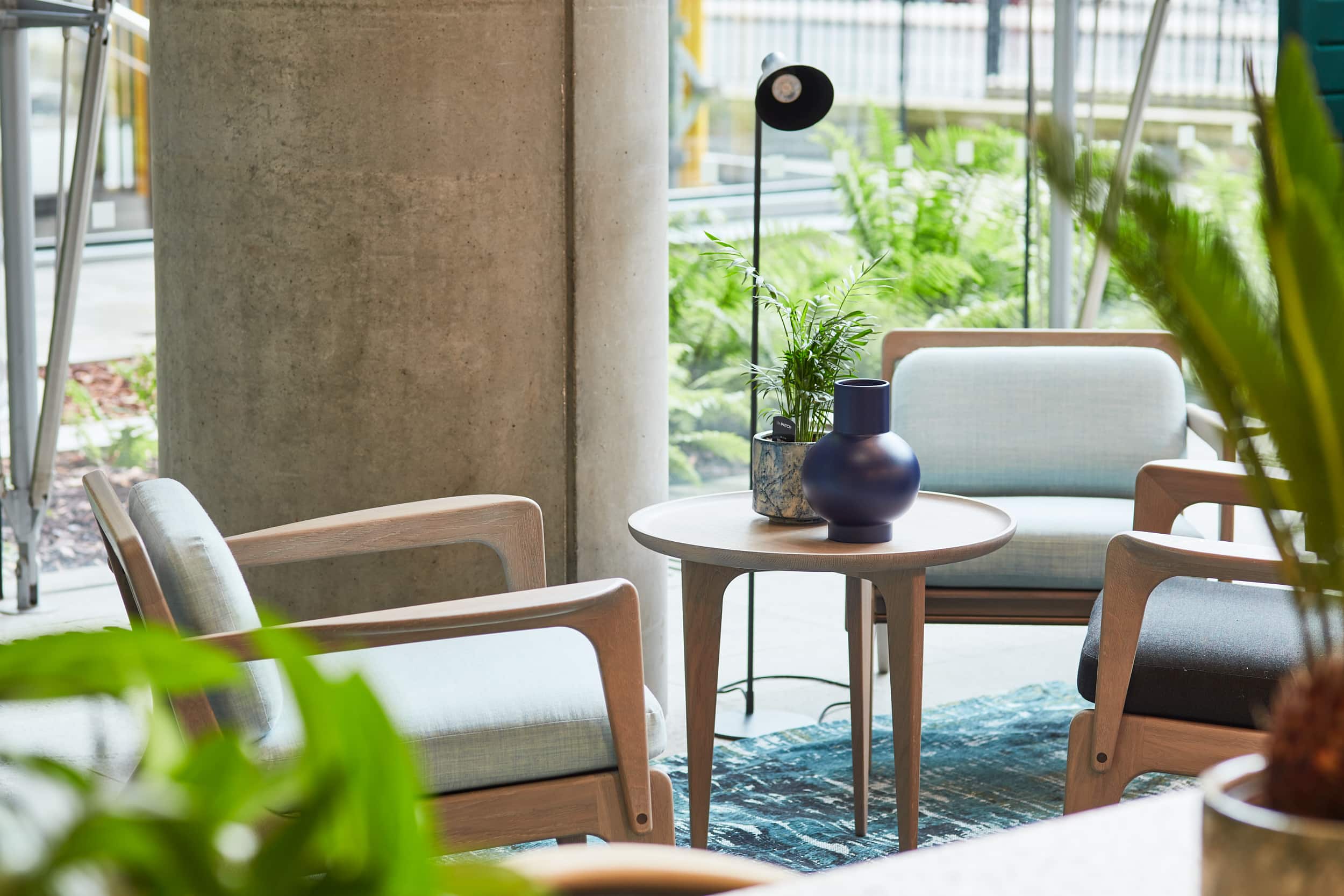 A cozy seating area featuring two wooden chairs with light blue cushions, a round wooden table with a dark blue vase and a small potted plant, surrounded by gre
