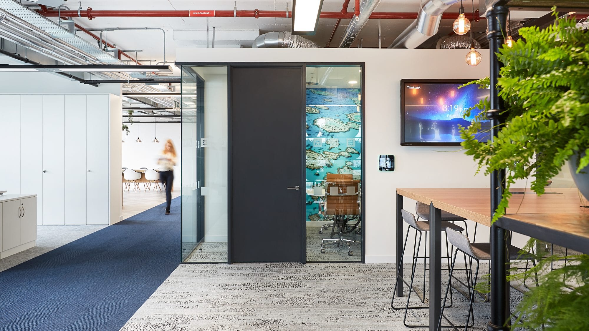 A women is walking down the hallway of a natural light filled workspace that also features lots of plants both exemplifying ways to improve your workplace wellbeing through office design.