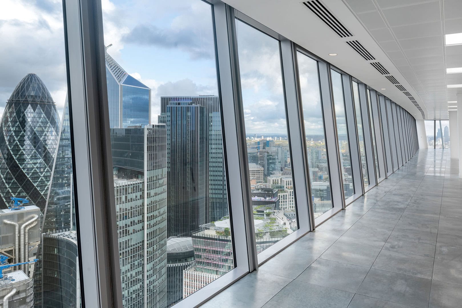 Interior view of a modern office space with large windows overlooking the London skyline, featuring various skyscrapers and cloudy skies.