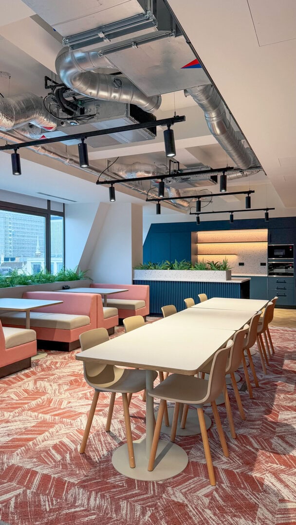 Modern office interior featuring a long table surrounded by chairs, pink seating areas, and a kitchen space with plants and industrial-style ceiling fixtures.
