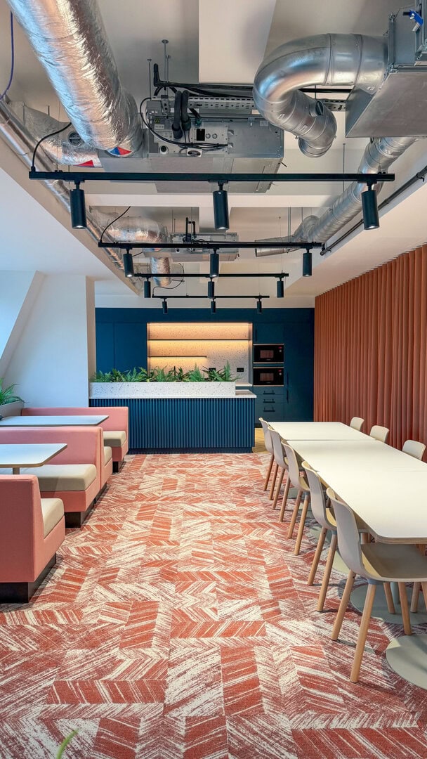 Modern office kitchen area featuring pink and gray seating, a long white table, and a blue cabinetry with plants, under exposed ductwork and track lighting.