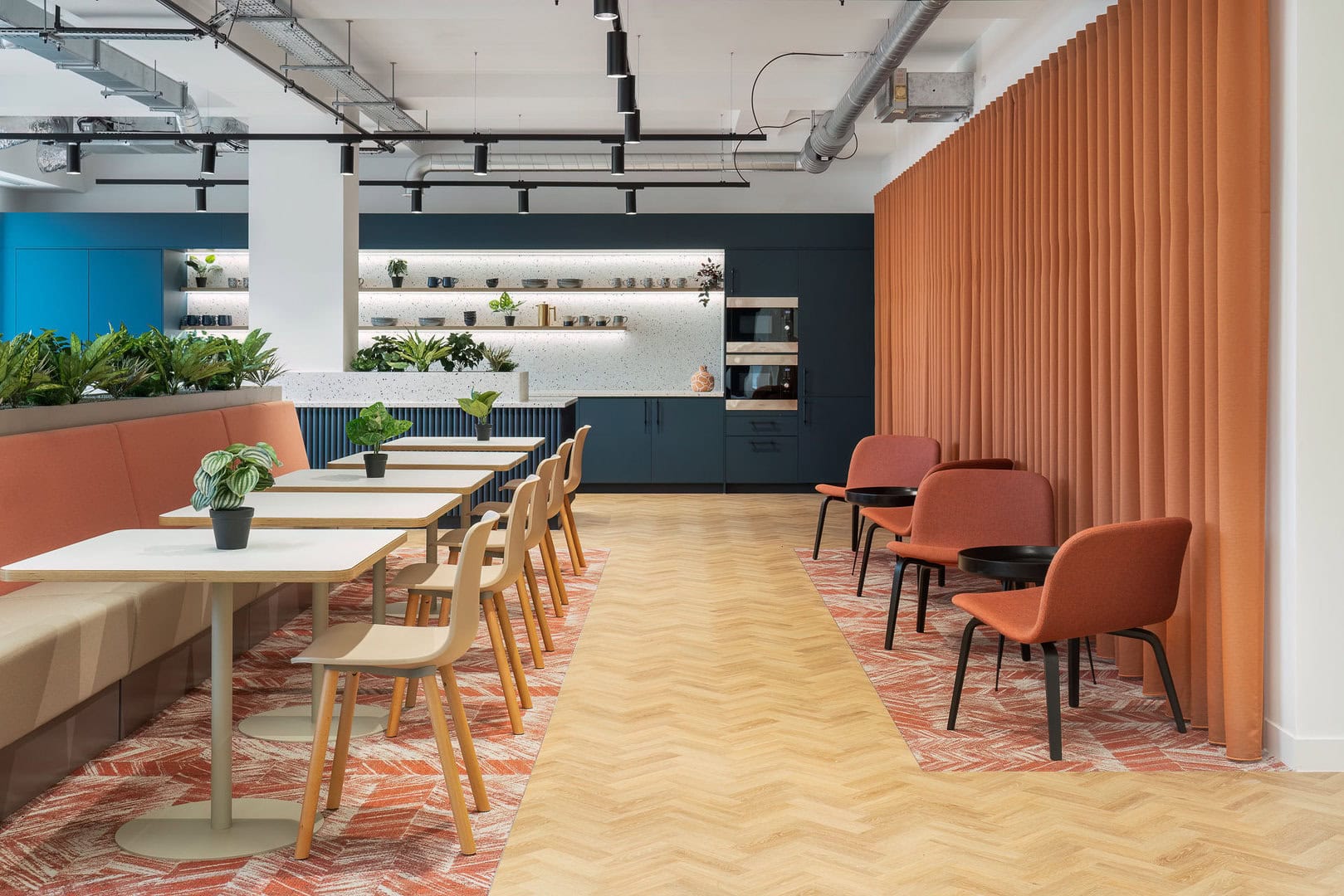 Modern workspace featuring a long table with chairs, a cozy seating area with orange curtains, and greenery, set against a backdrop of blue cabinetry and a ligh