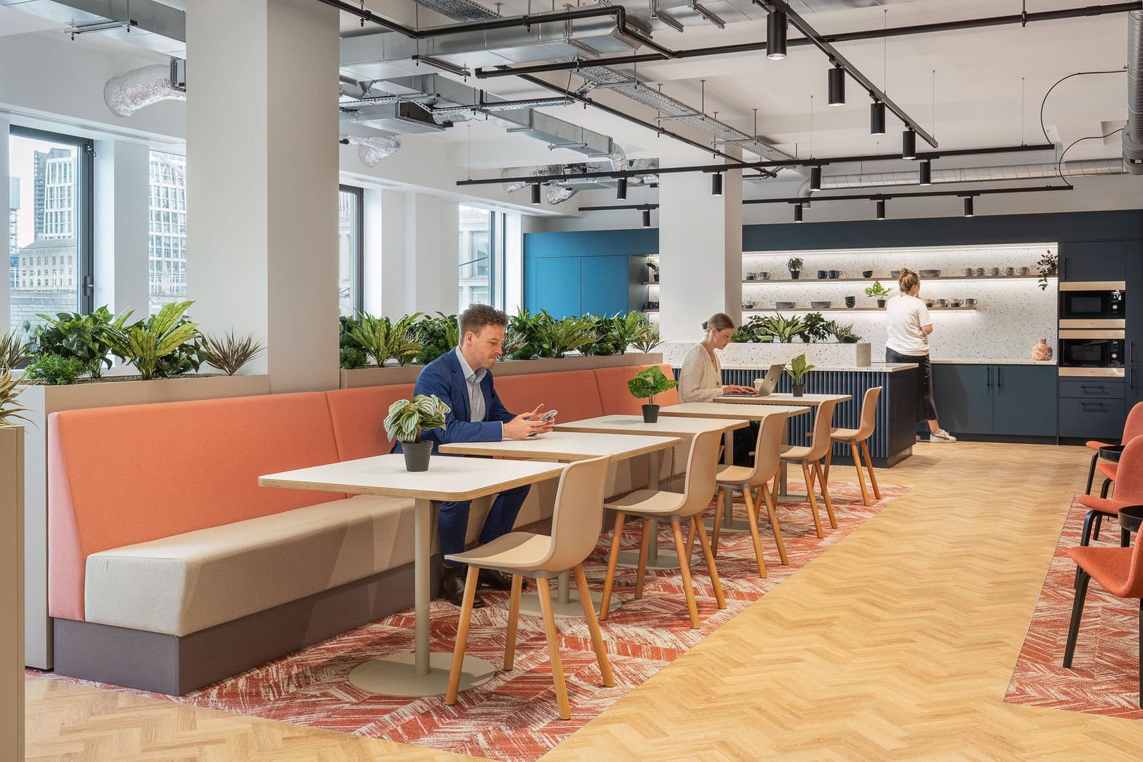 Modern office interior featuring a seating area with a man using a smartphone, a woman working on a laptop, and greenery throughout the space.