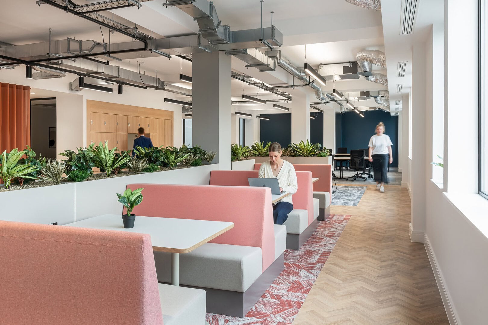 Modern office interior featuring pink and gray seating areas, greenery, and individuals working on laptops.
