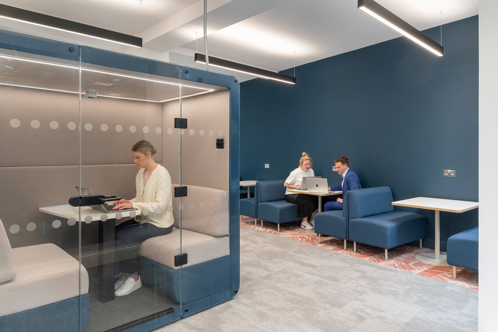 A modern workspace featuring a glass-enclosed booth with a woman using a laptop, and a seating area where a man and woman are collaborating on a laptop.