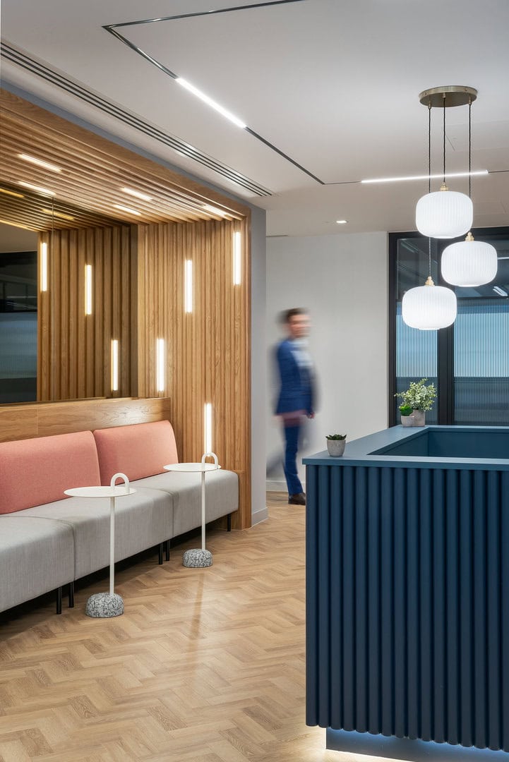 Modern reception area featuring a blue front desk, pink and gray seating, wooden wall accents, and pendant lighting, with a blurred figure walking in the backgr
