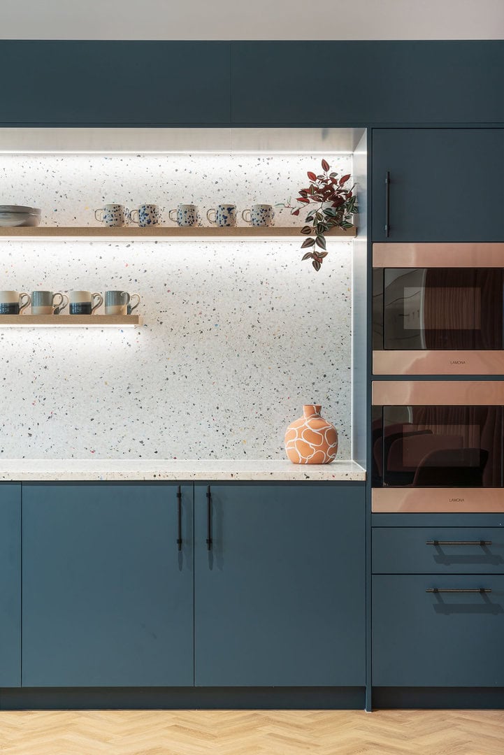 Modern kitchen featuring blue cabinetry, a speckled countertop, decorative mugs on a wooden shelf, and an orange patterned vase.