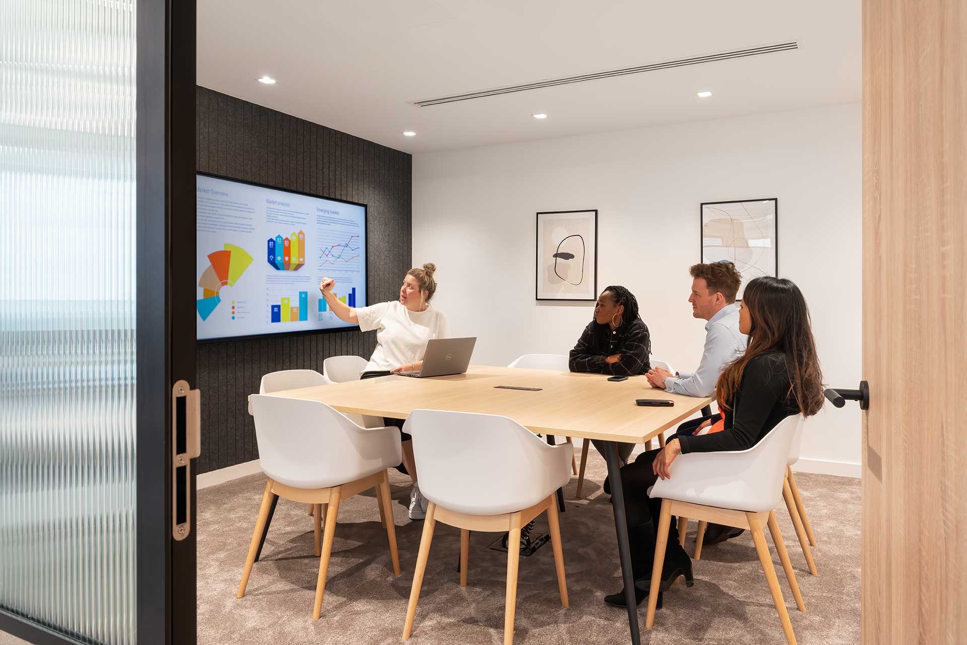 A woman presents data on a screen to a group seated around a conference table in a modern meeting room.