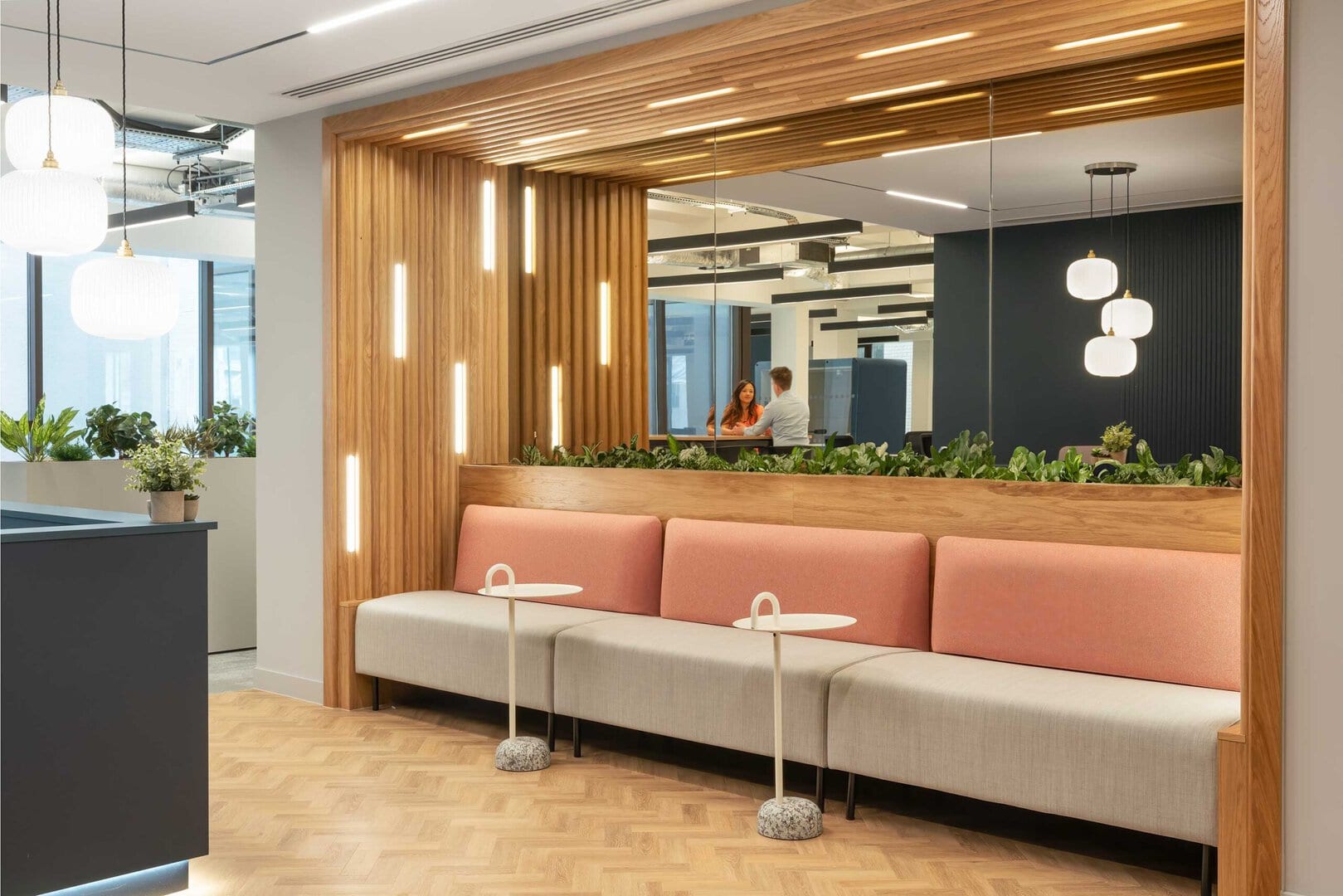 Modern office lounge area featuring a wooden accent wall, pink and gray seating, small white tables, and decorative plants, with two people visible in the backg