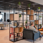 Two men are seated in modern lounge chairs, engaged in conversation, with a backdrop of a contemporary office space featuring plants and a glass meeting room.