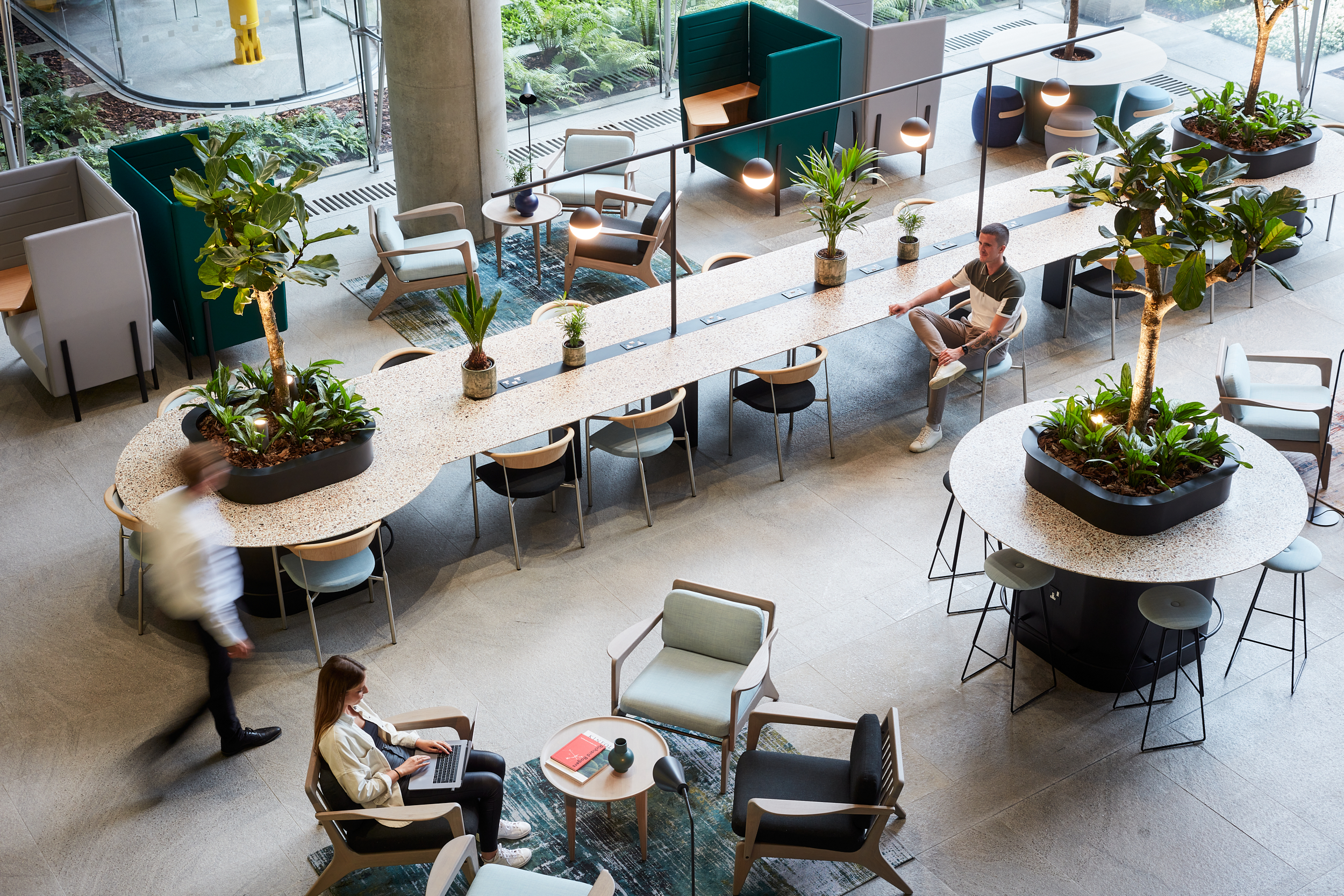 A modern workspace featuring a long table with potted plants, surrounded by various seating options, with a person working on a laptop and another walking in th