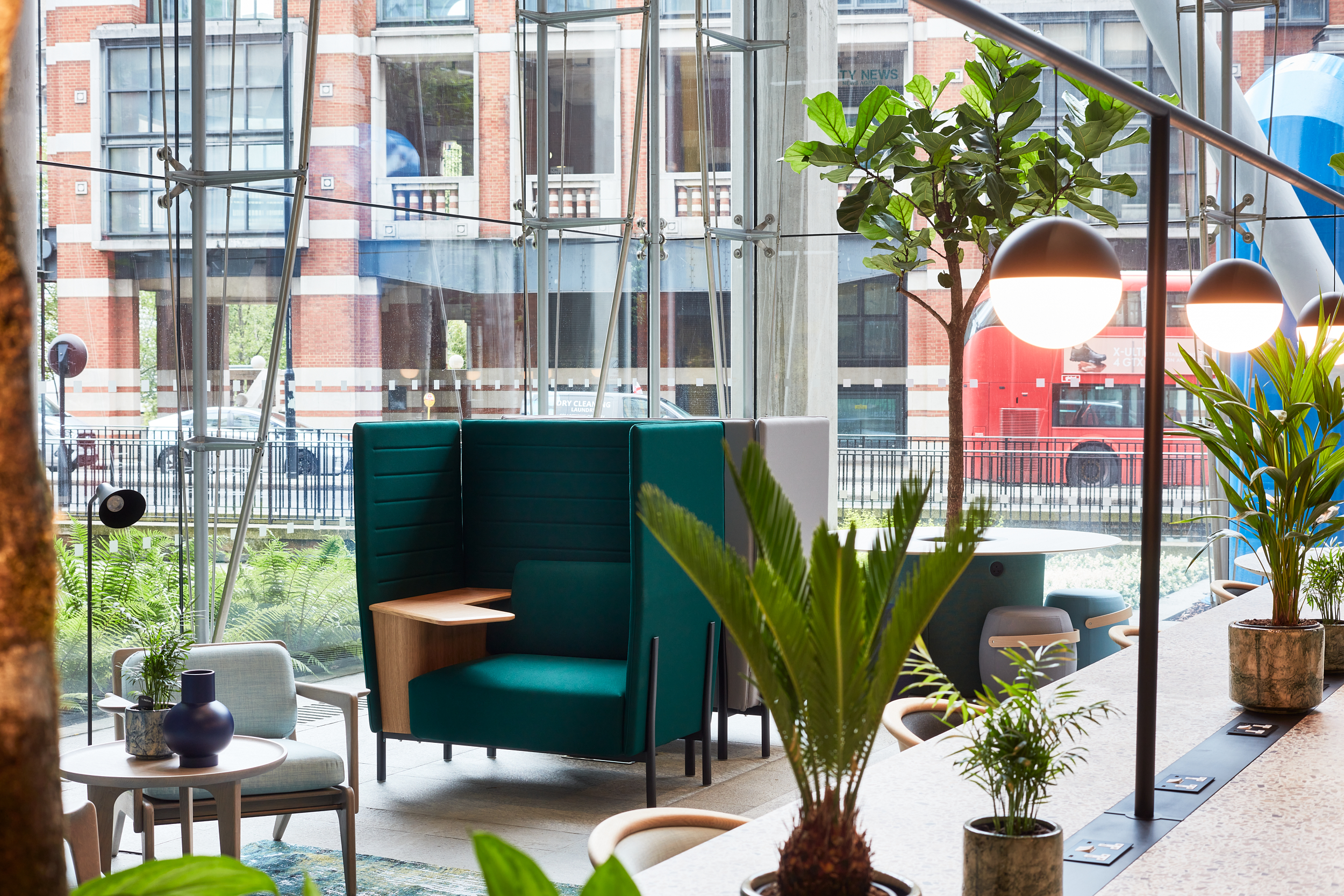 Interior view of a modern seating area featuring a green upholstered booth, wooden side table, and various indoor plants, with large windows showing a city stre