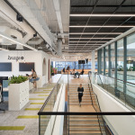 Modern office interior featuring open workspaces, greenery, and large windows, with people engaged in various activities and a staircase in the foreground.