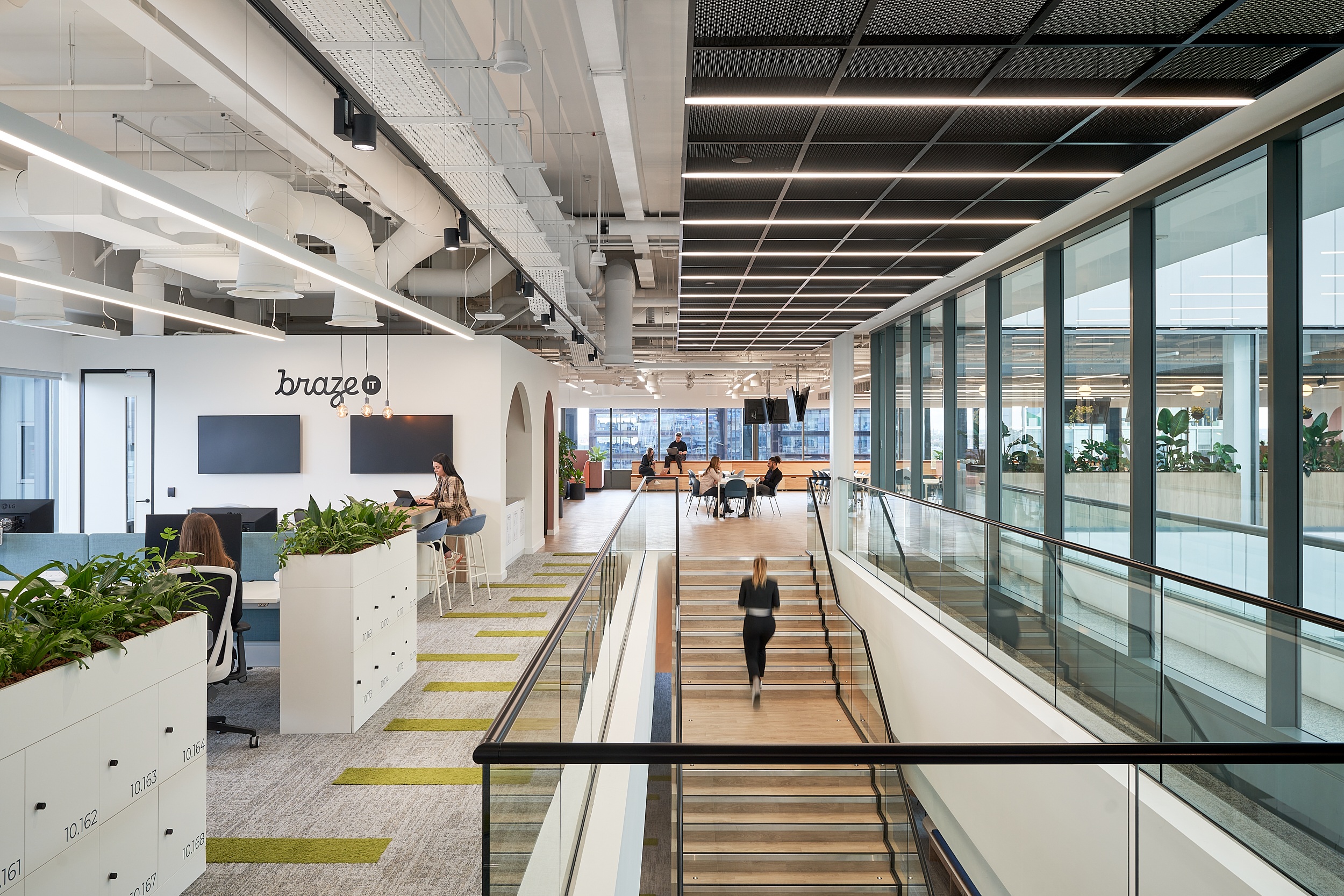 Modern office interior featuring open workspaces, greenery, and large windows, with people engaged in various activities and a staircase in the foreground.