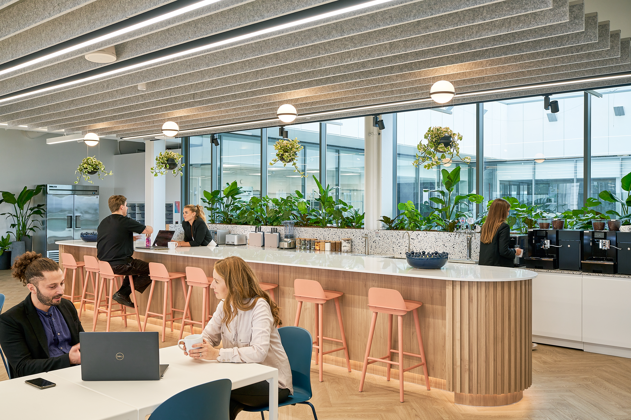 A modern office kitchen with a long countertop, pink bar stools, and large windows filled with greenery, featuring two people at the counter and two others seat
