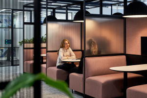 A woman sits at a table in a modern office booth, engaged in conversation with another person whose face is obscured by a frosted partition.