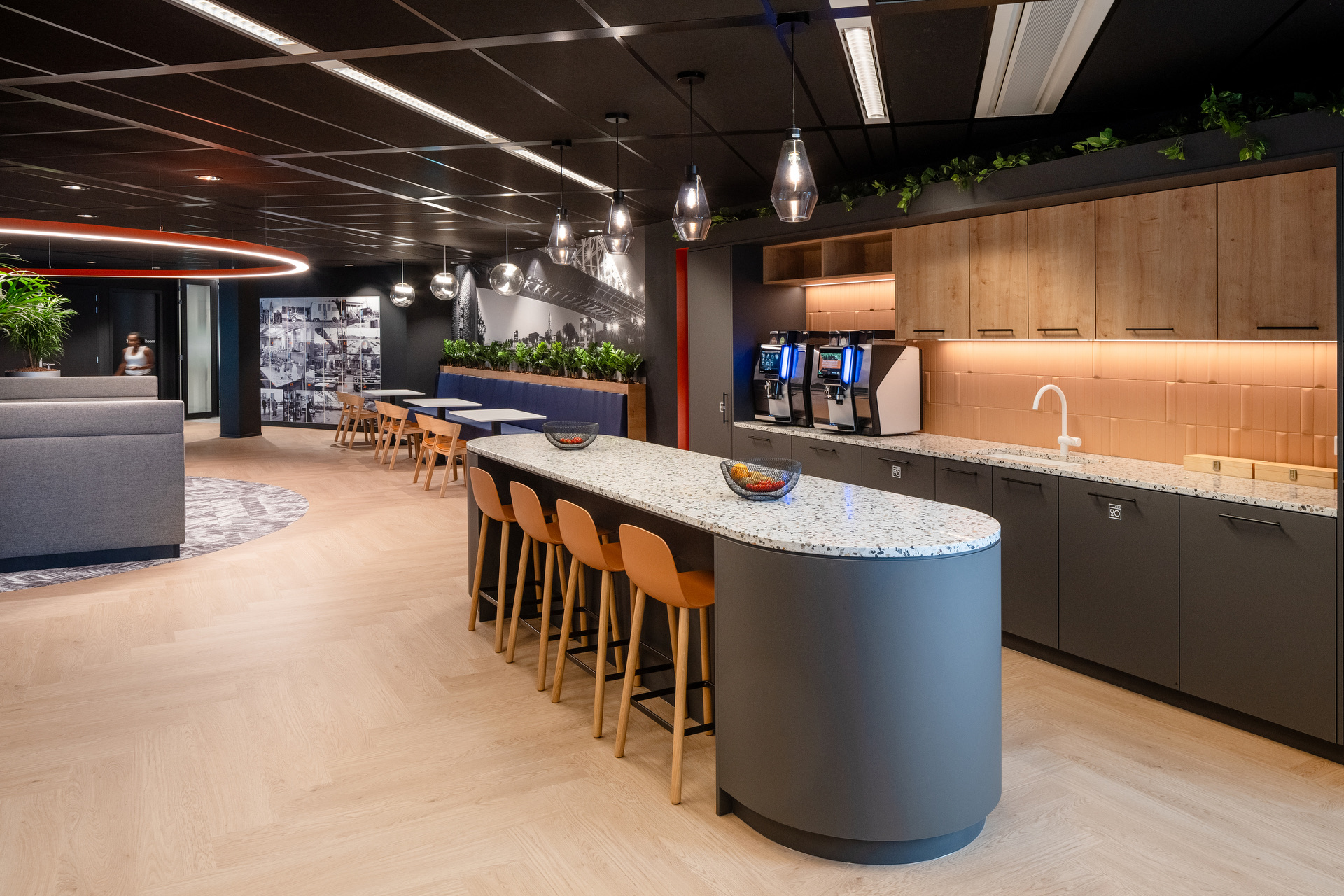Modern office kitchen area featuring a curved granite countertop with seating, coffee machines, wooden cabinetry, and greenery.