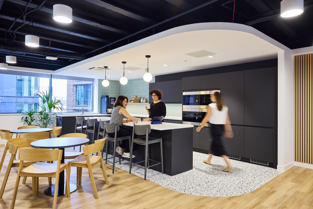 Modern office kitchen with two women interacting at a counter, bar stools, and a dining area featuring wooden tables and chairs, large windows, and contemporary