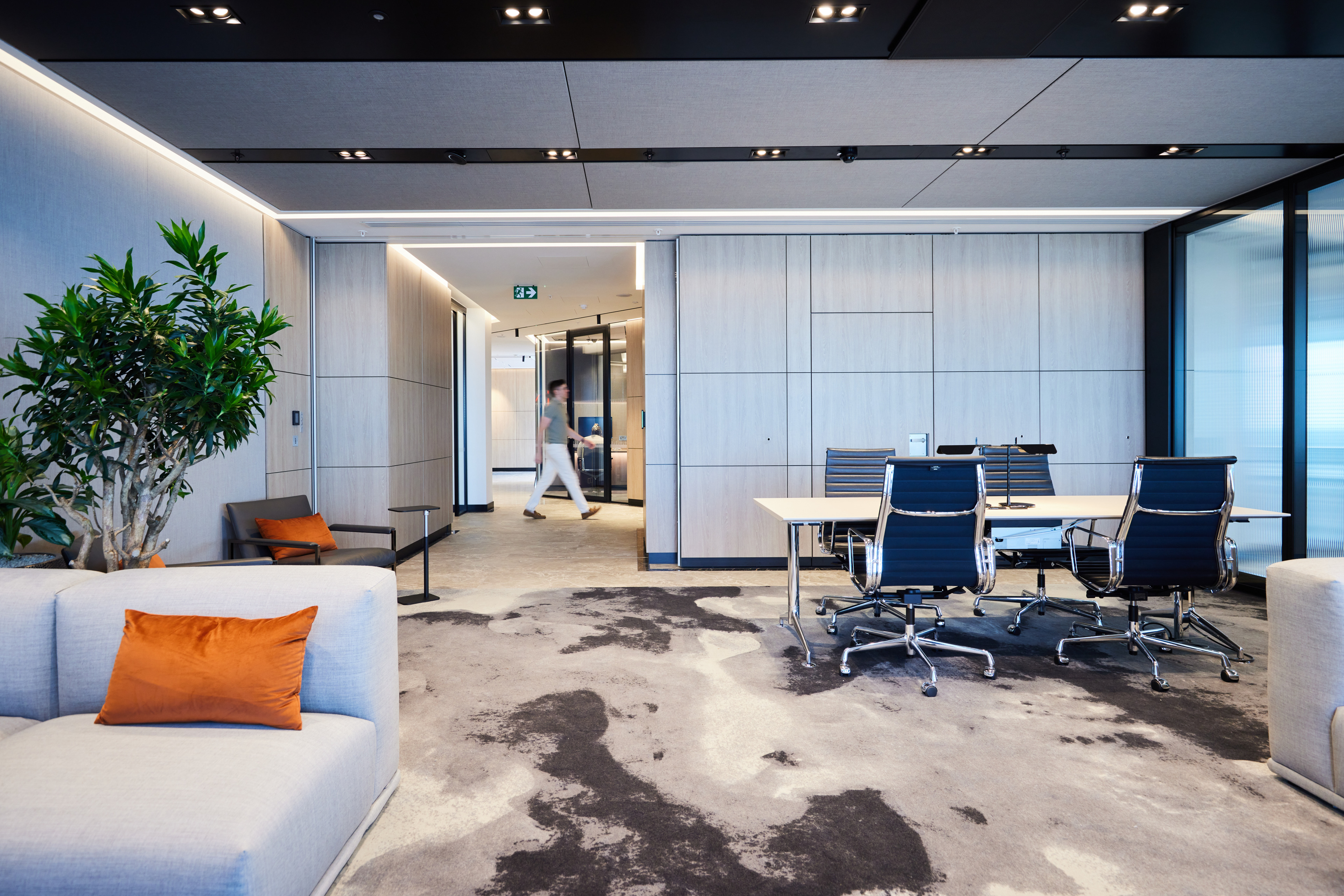 Modern office interior featuring a seating area with a gray couch and orange pillow, a potted plant, and a conference table with black chairs, with a person wal
