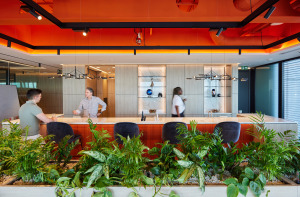 Modern office interior featuring a bar area with two men engaged in conversation, surrounded by greenery and contemporary decor.