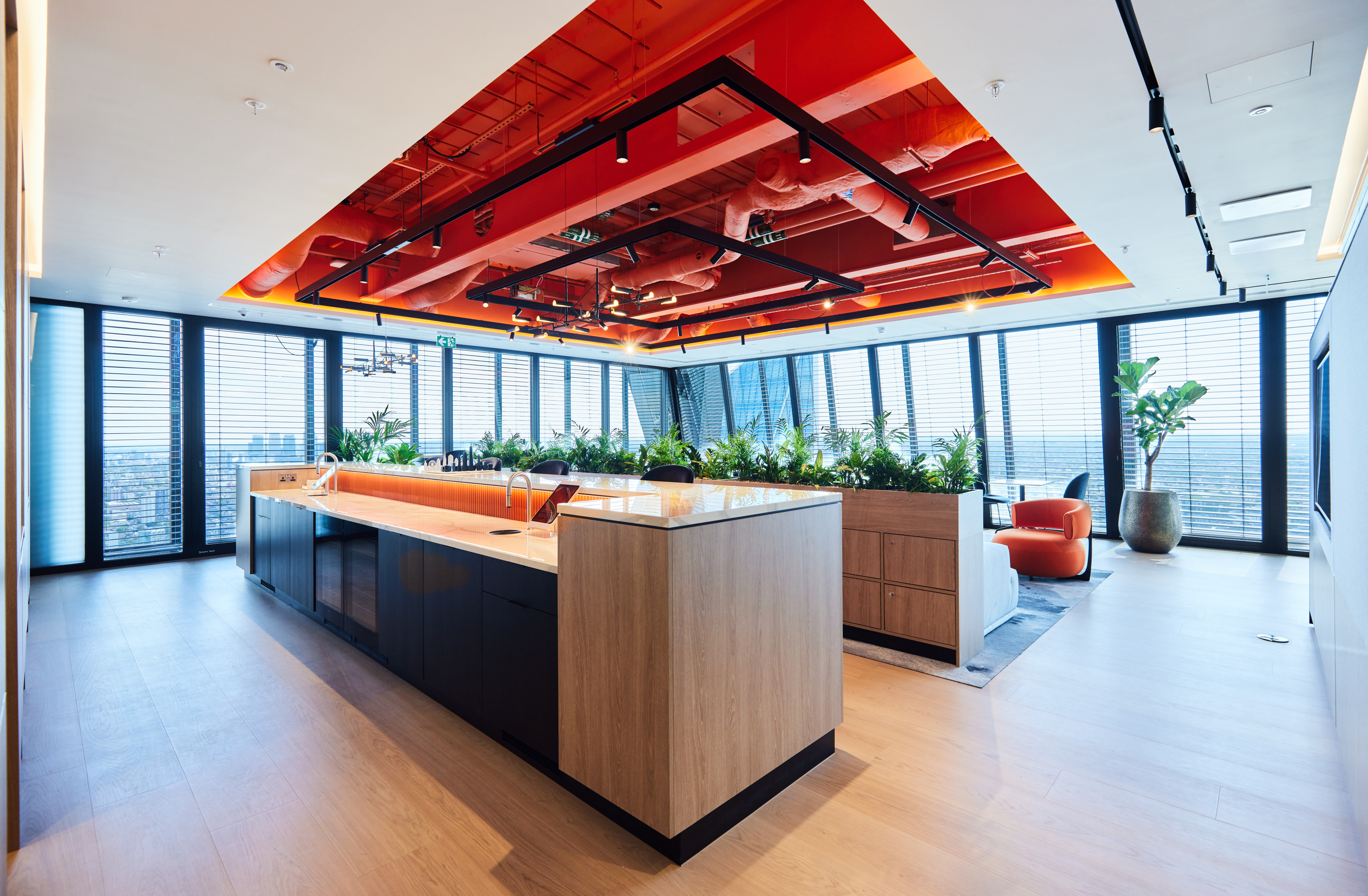Modern office interior featuring a kitchen area with a large countertop, wooden cabinetry, and lush greenery, alongside large windows offering a city view.