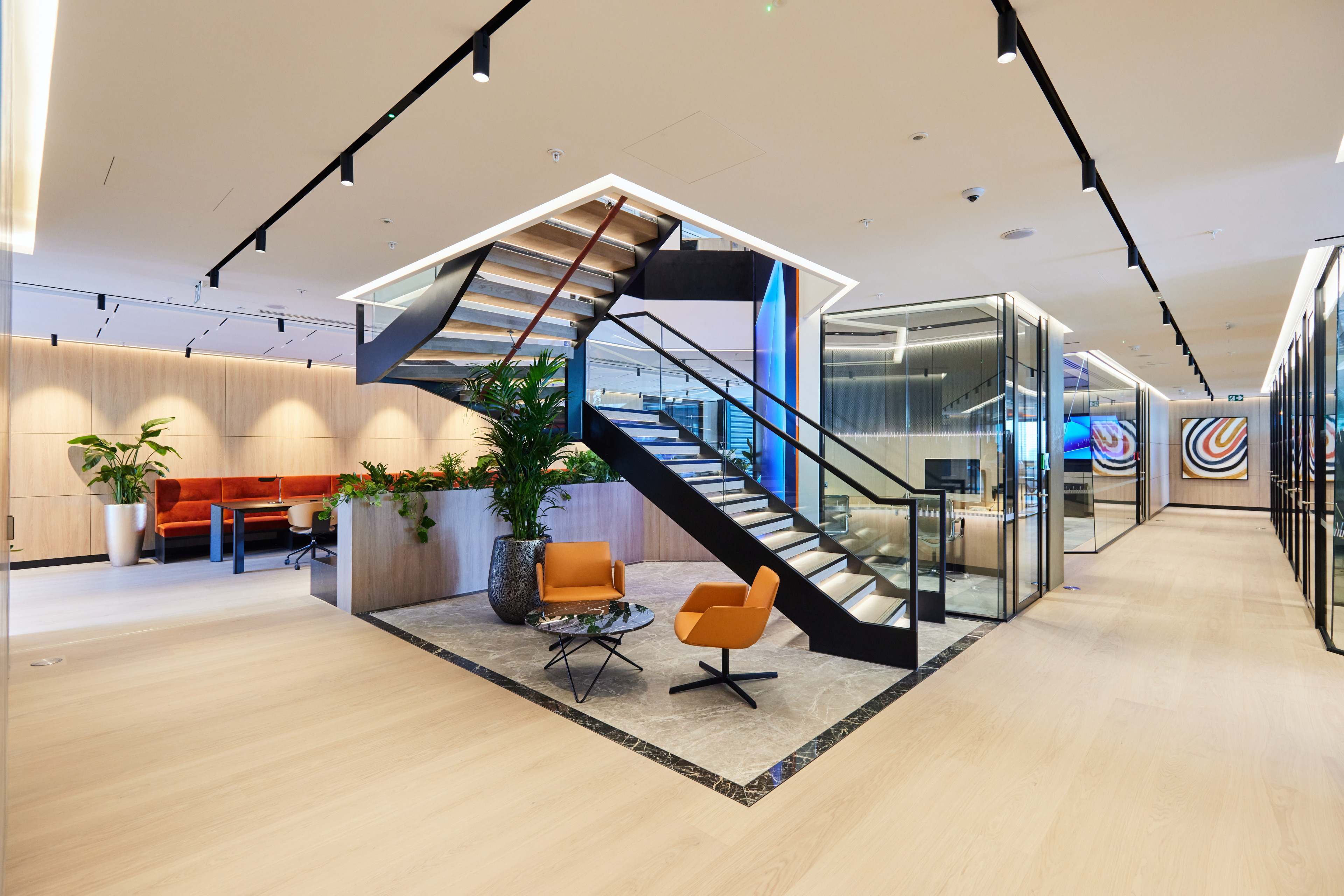 Modern office interior featuring a staircase, seating area with orange chairs, and glass-walled meeting rooms.