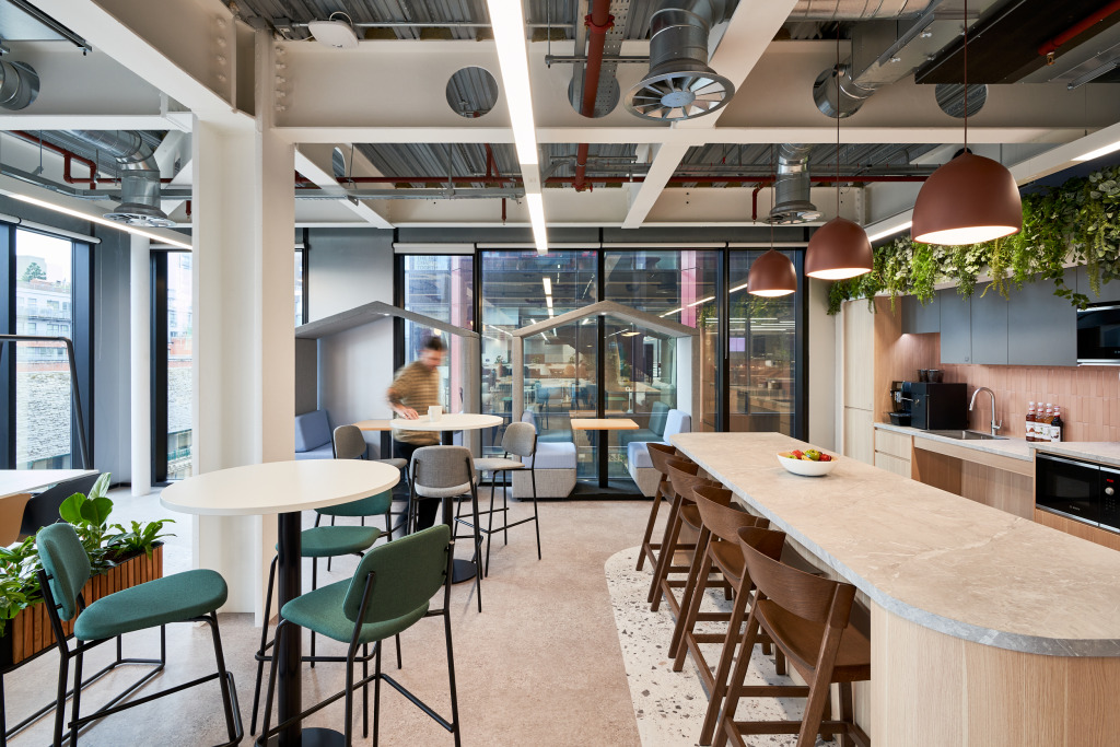 Modern office kitchen area featuring a long countertop, bar stools, and tables, with large windows and greenery, and a person in motion.