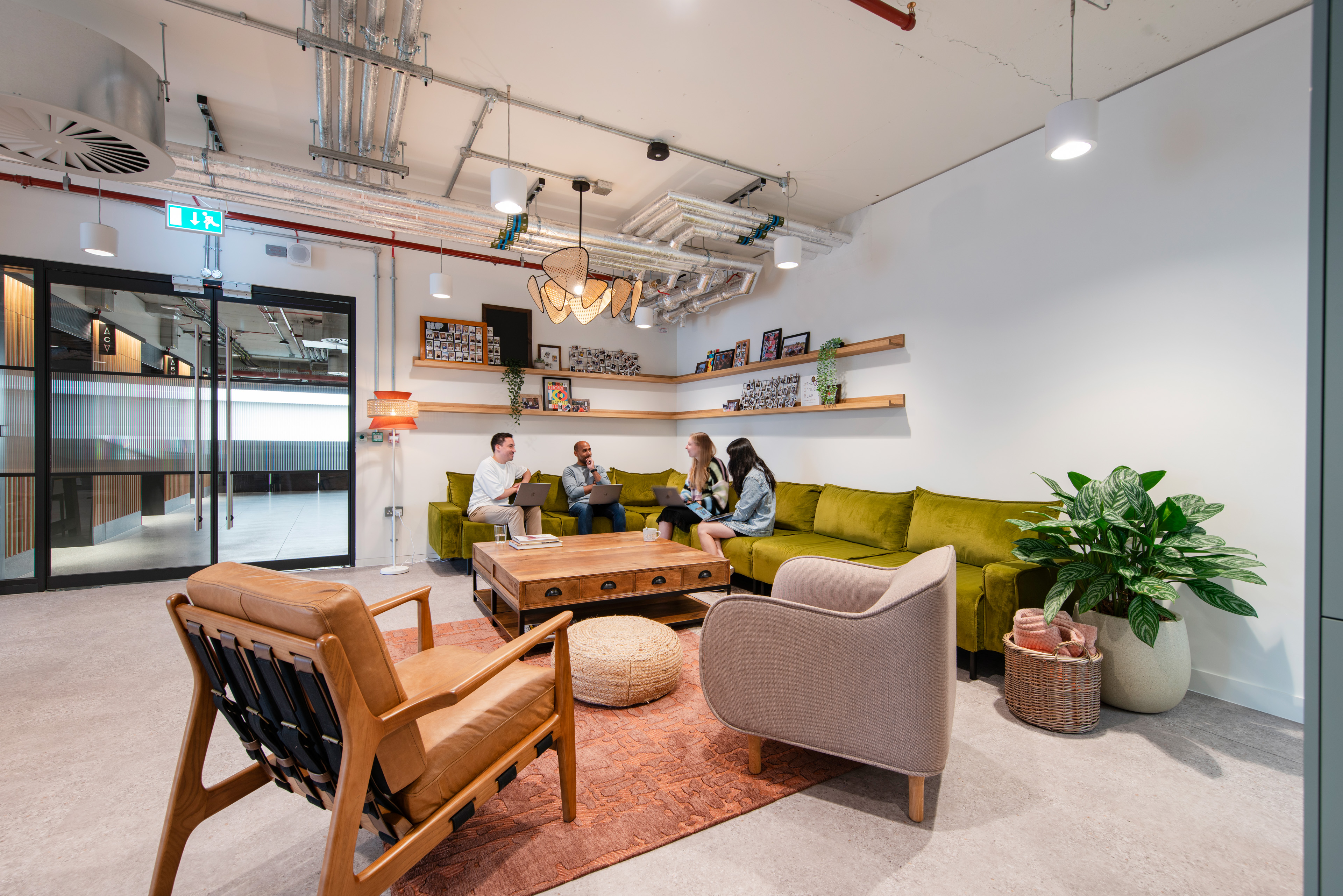 A modern lounge area featuring a green sectional sofa, wooden coffee table, and various plants, with four people engaged in conversation and using laptops.