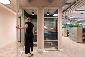 A person in black clothing walks through a glass door into a modern office space with green plants and workstations.