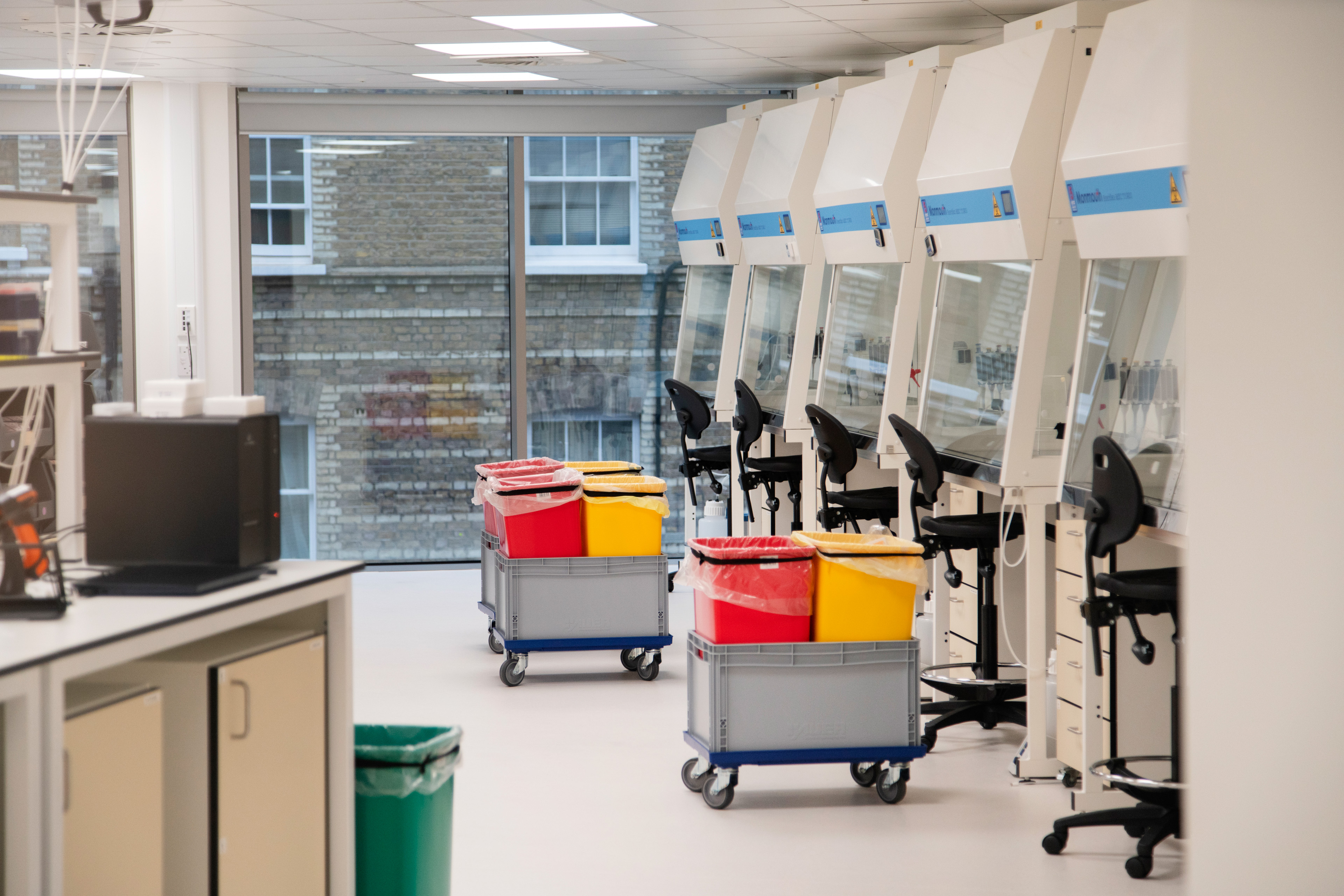 Laboratory interior featuring several biosafety cabinets, rolling carts with color-coded waste bins, and a view of brick buildings through large windows.